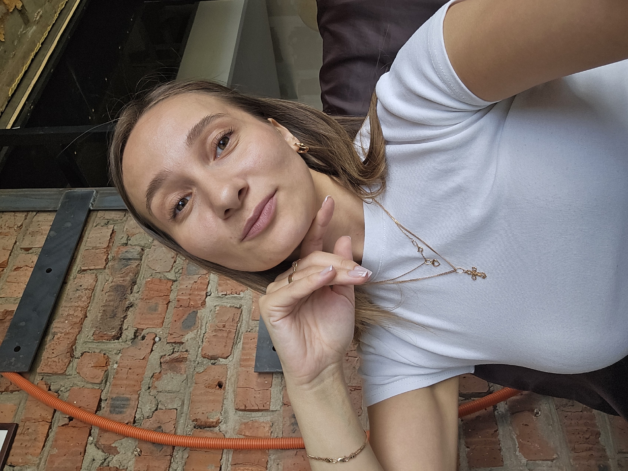 A woman takes a selfie. She is wearing a white t-shirt and gold jewelry, including a necklace with a cross pendant. She is leaning against a brick wall. An orange corrugated tube is visible near the bottom of the frame. The location is Chaikovsky, Russia.