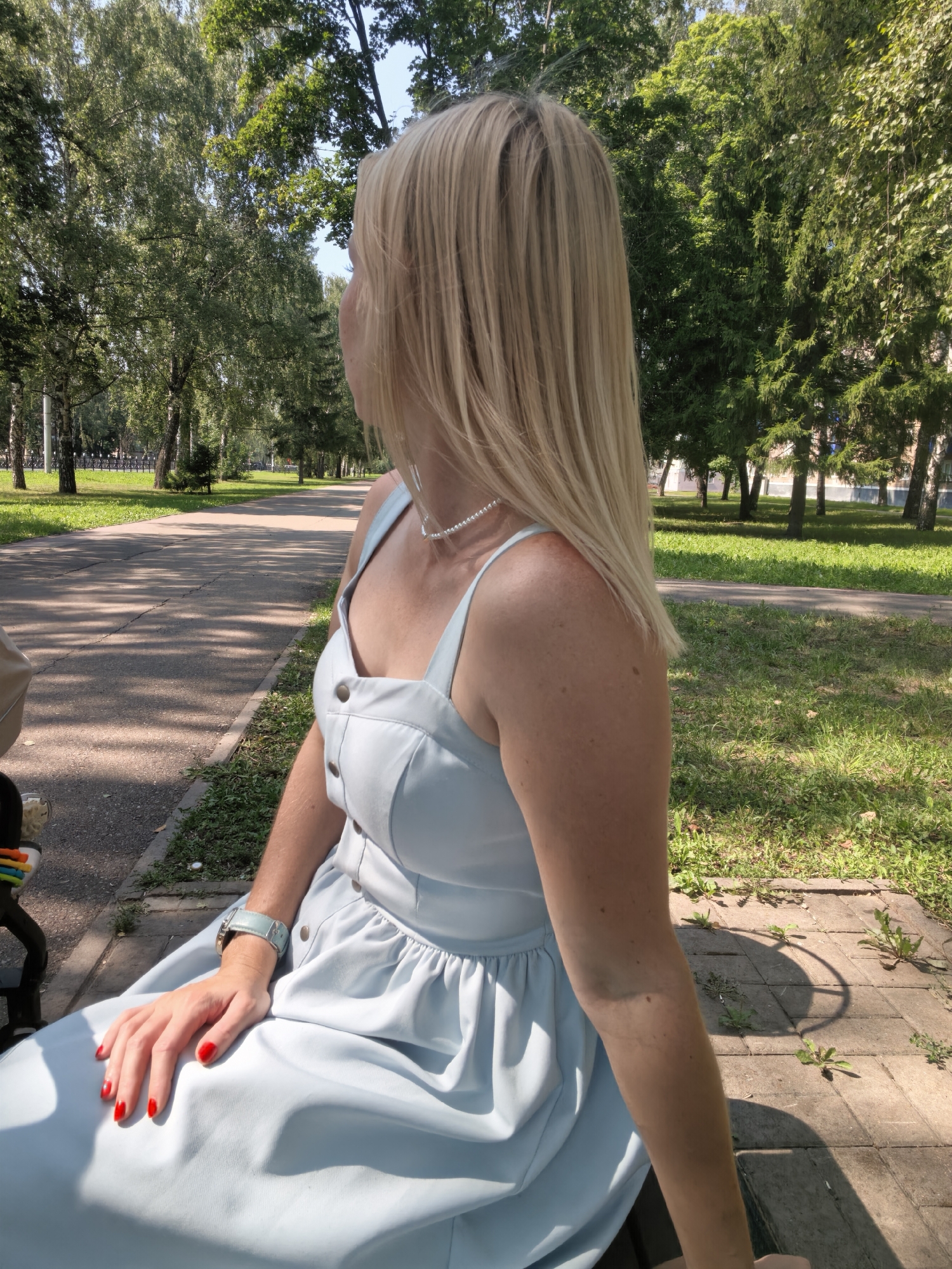 A medium shot shows a woman with blonde hair, wearing a light blue dress and a pearl necklace, sitting on a bench in a park. Her face is turned away from the camera, looking towards the left. The park has a paved walkway and is lined with trees and green grass. The woman's right hand, with red nail polish, rests on her lap. A light blue watch is visible on her left wrist. The image is taken on a sunny day, with shadows cast by the trees on the ground. The location is not explicitly identifiable as Sterlitamak, Russia, based solely on the visual information provided.