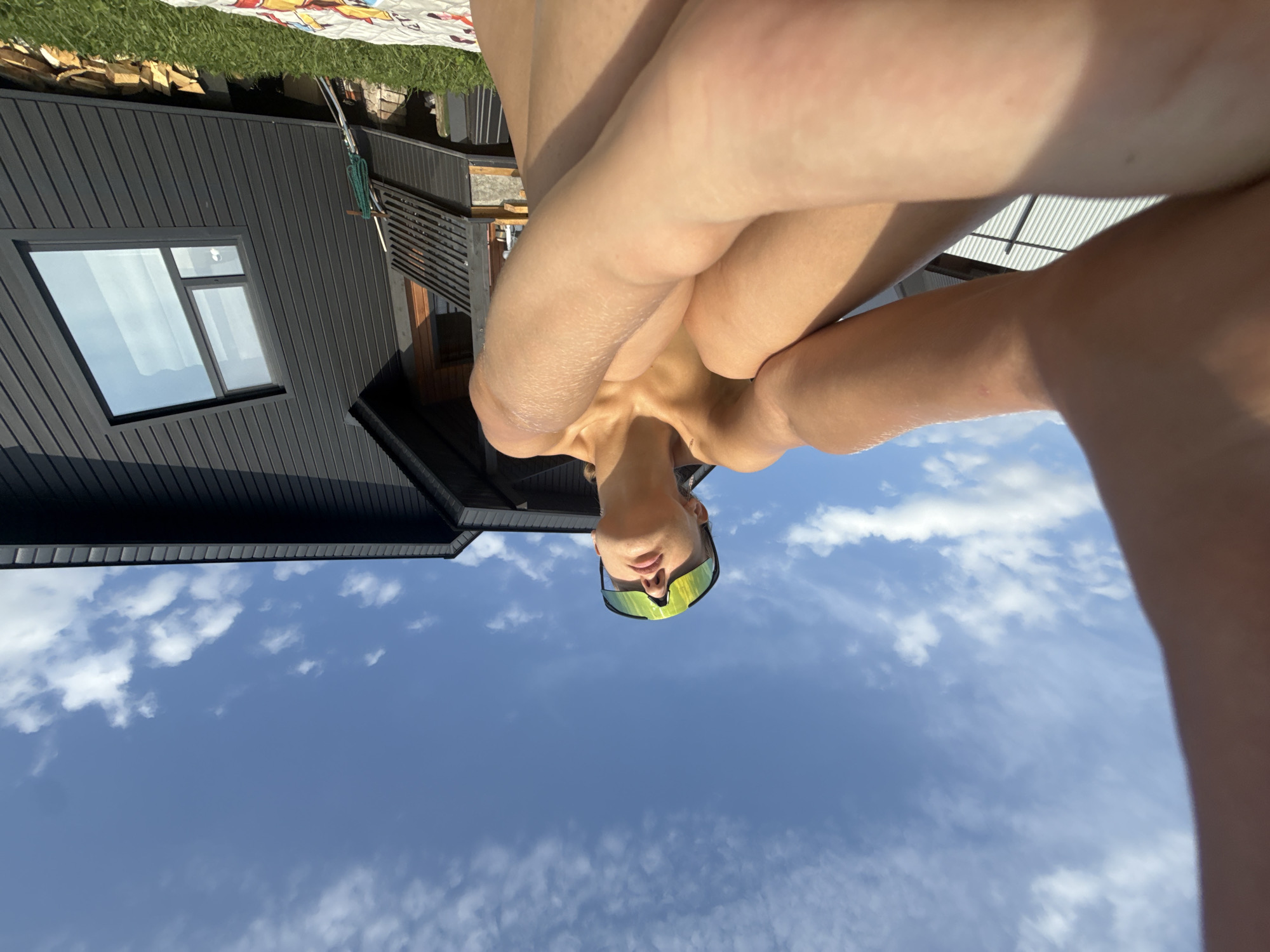 A person is sitting outside in a crouched position on a patterned blanket, facing upwards towards a blue sky with scattered clouds. They are wearing large, reflective sunglasses with a neon yellow tint. Their skin is tanned, and their arms are wrapped around their legs. In the background, a modern, dark-colored wooden house with a sloped roof and a large, angled window is visible. The house has a wooden balcony with horizontal railings. To the right of the house, a pile of chopped firewood rests on the grass. The overall environment suggests a sunny day outdoors. The location is identified as Podshivalovo, Russia.