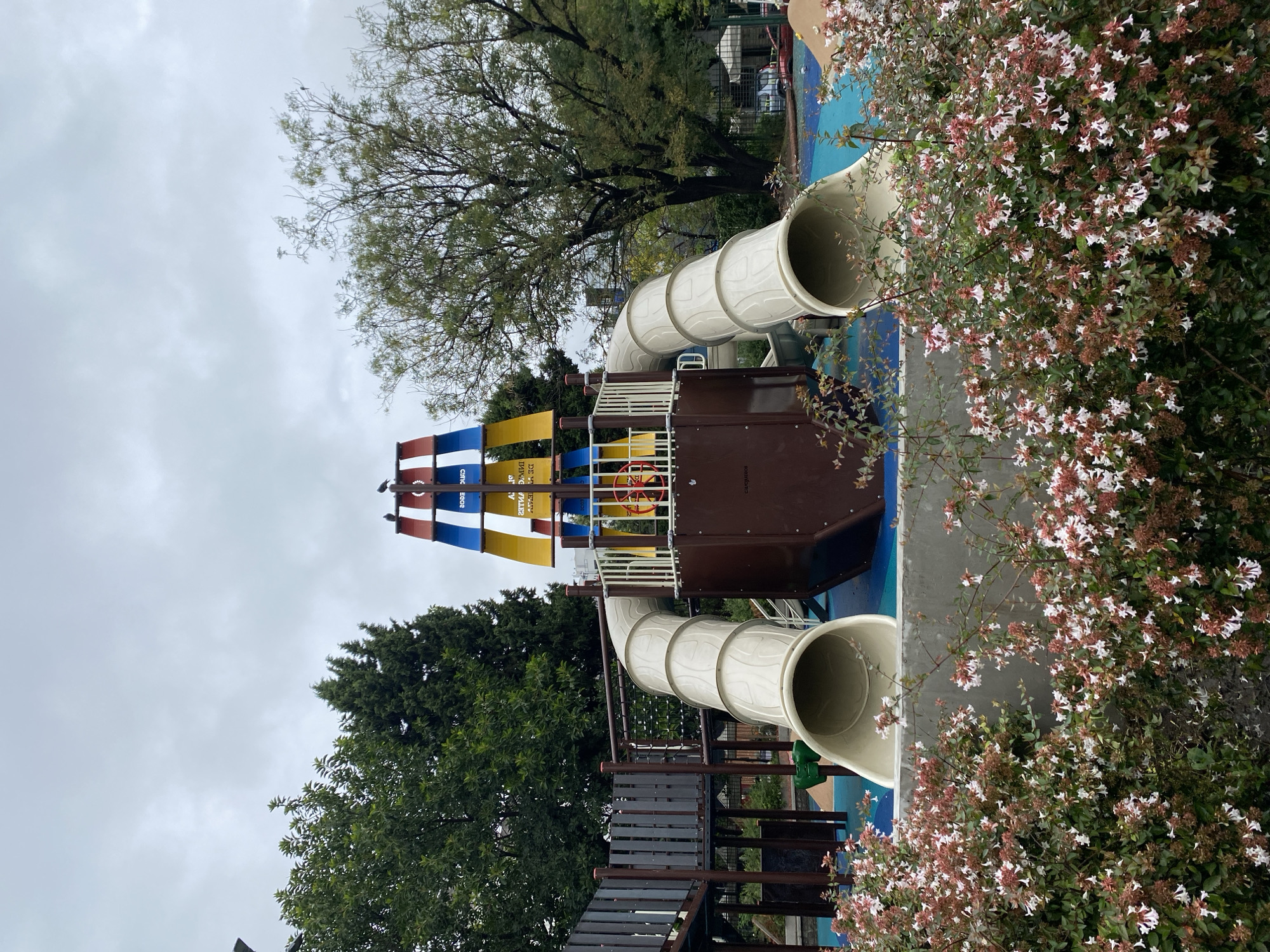 The scene is a playground in Buenos Aires, Argentina. It's daytime, under an overcast sky. The main structure is a pirate ship-themed play set with two enclosed beige slides spiraling down from a brown central platform. The ship's mast is decorated with blue, white, red, and yellow banners. The play area's ground is painted blue. A staircase made of dark wood leads to the play set. Trees are visible in the background. A bush with white and red flowers grows beside the play set.