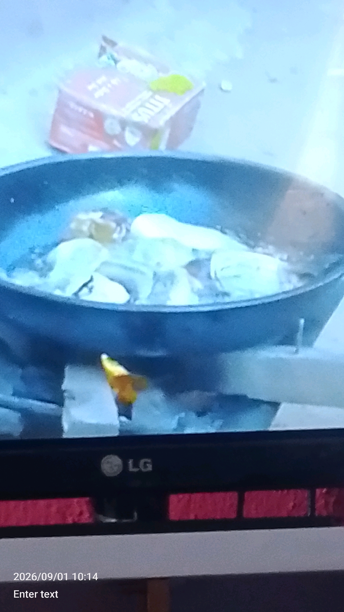 The image shows a close-up of a frying pan filled with food, possibly plantains, being cooked over an open flame. The pan is dark and appears to be made of cast iron. The flame is visible underneath, producing smoke and heat. In the background, a partially visible cardboard box with some writing on it can be seen. The overall scene suggests an outdoor cooking setup, possibly in a residential area of Lokoja, Nigeria. The bottom of the image displays a portion of an LG television screen, with a date and time stamp indicating "2026/09/01 10:14" and the text "Enter text". The presence of the television suggests the image may have been captured from a screen displaying a live feed or recording.