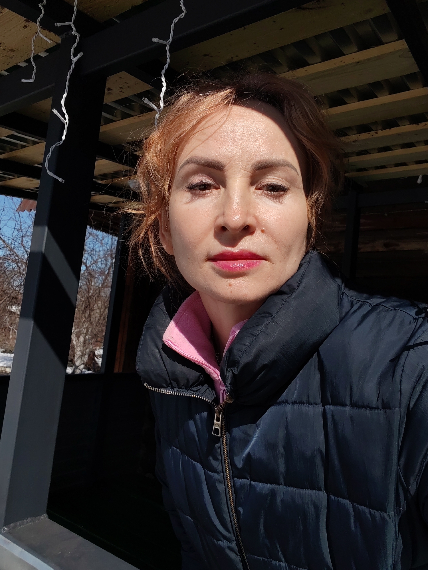 A woman is centered in the frame, looking directly at the viewer. She has reddish-blonde hair pulled back from her face and is wearing a navy blue puffer jacket with a pink collar visible beneath. She is positioned within a gazebo-like structure with wooden planks overhead and black metal supports visible on either side. Through an opening in the structure, trees without leaves are seen against a blue sky. Remnants of snow are visible on the ground in the distance. String lights are visible hanging from the ceiling of the structure.
The location is Chaikovsky, Russia.