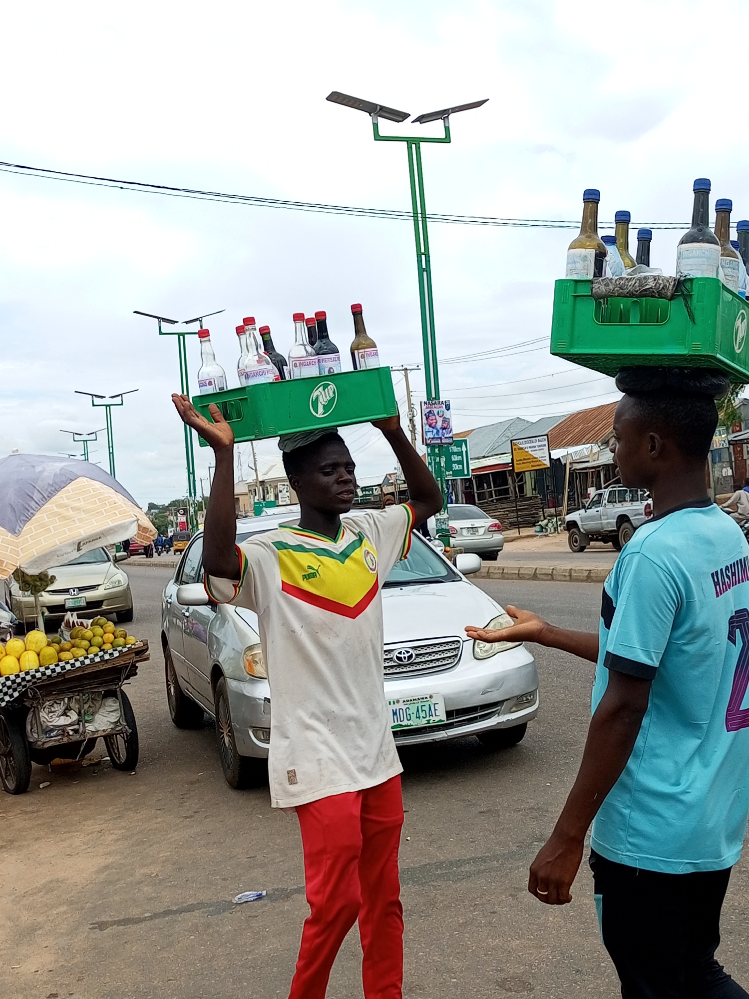 This media file shows two young men in a street scene in Tempure, Nigeria. The man on the left, wearing a white jersey with green and yellow accents and red pants, carries a green plastic crate filled with glass bottles on his head. He is looking to his right, with his left arm raised. The man on the right, wearing a light blue t-shirt with black accents and black pants, is facing the man on the left. His left arm is extended, and he appears to be speaking or gesturing.
In the background, several cars are visible on a paved road, along with streetlights and buildings. To the left of the main subjects, there is a fruit cart laden with yellow and orange fruits, covered by a patterned umbrella. The sky is overcast, suggesting a cloudy day. The overall atmosphere appears to be that of a busy street market or roadside commerce.
Visible text includes "LAPARGE" on the umbrella, and labels on the bottles such as "INGANCHI FE" and "INGANCY HERBAL". A license plate on a silver car reads "MDG-45AE ADAMAWA". Text on a signpost indicates distances: "176km", "60km", and "50km". The back of the blue shirt worn by the man on the right has the name "HASHIM" visible.
