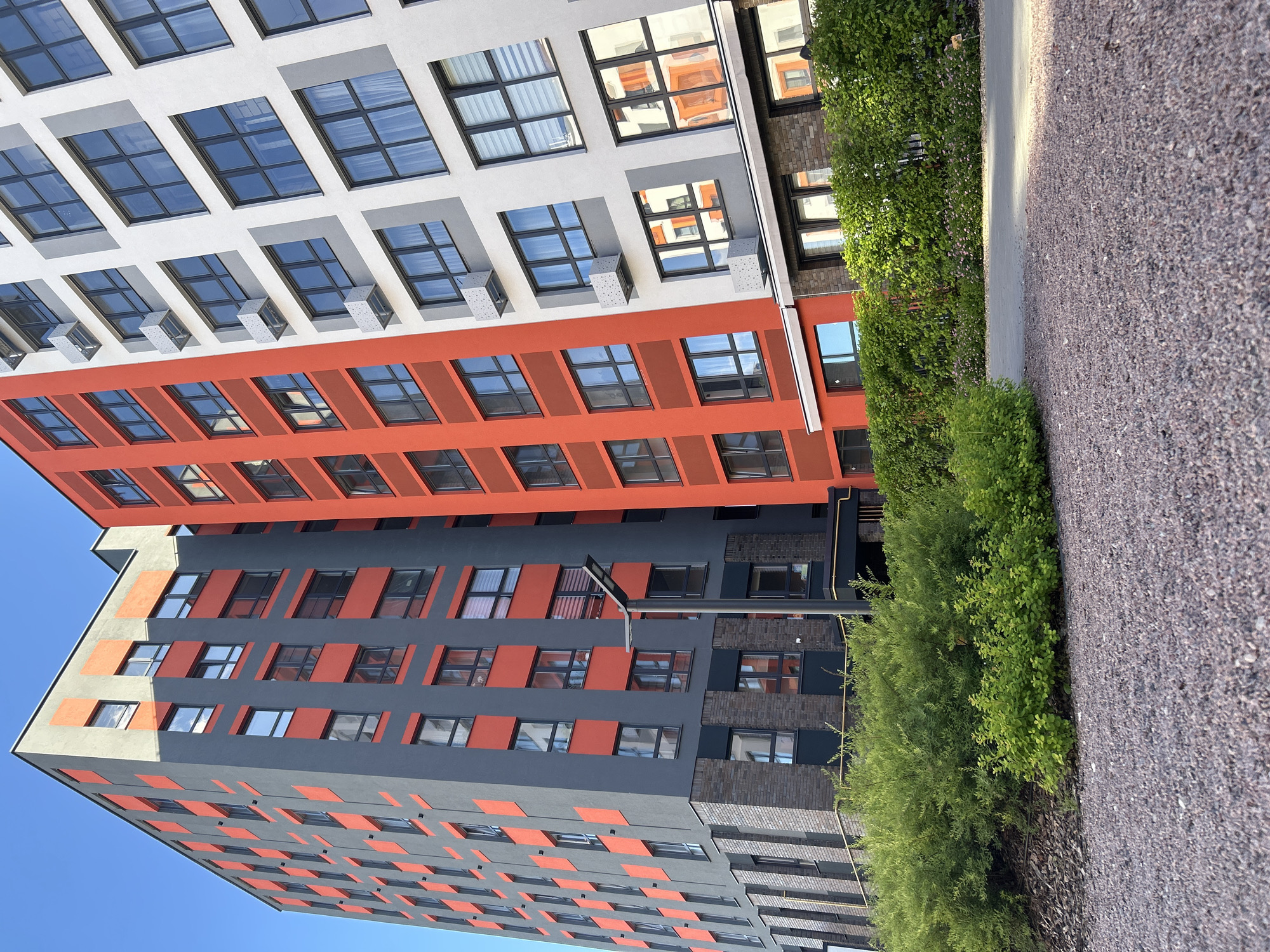 This is a vertical, medium shot of a modern residential building in Kaluga, Russia. The building features a striking architectural design with alternating sections of white, bright orange, and dark gray facades. Numerous square windows are visible on each level, many with dark frames, some reflecting the surrounding environment. The building's structure extends upwards and appears to be an apartment complex.
In the foreground, a narrow pathway composed of gravel runs alongside a dense line of lush green bushes and trees. The foliage is vibrant and appears to be well-maintained, suggesting a park-like or landscaped area around the building. The sky is a clear, bright blue, indicating fair weather and likely daytime, possibly late morning or early afternoon given the shadows cast by the building.
There is no visible text in the image. The scene depicts a quiet, modern urban setting, likely a residential area designed with contemporary aesthetics. The overall impression is one of clean lines, bright colors, and well-kept greenery. The image does not capture any people or specific activities.
