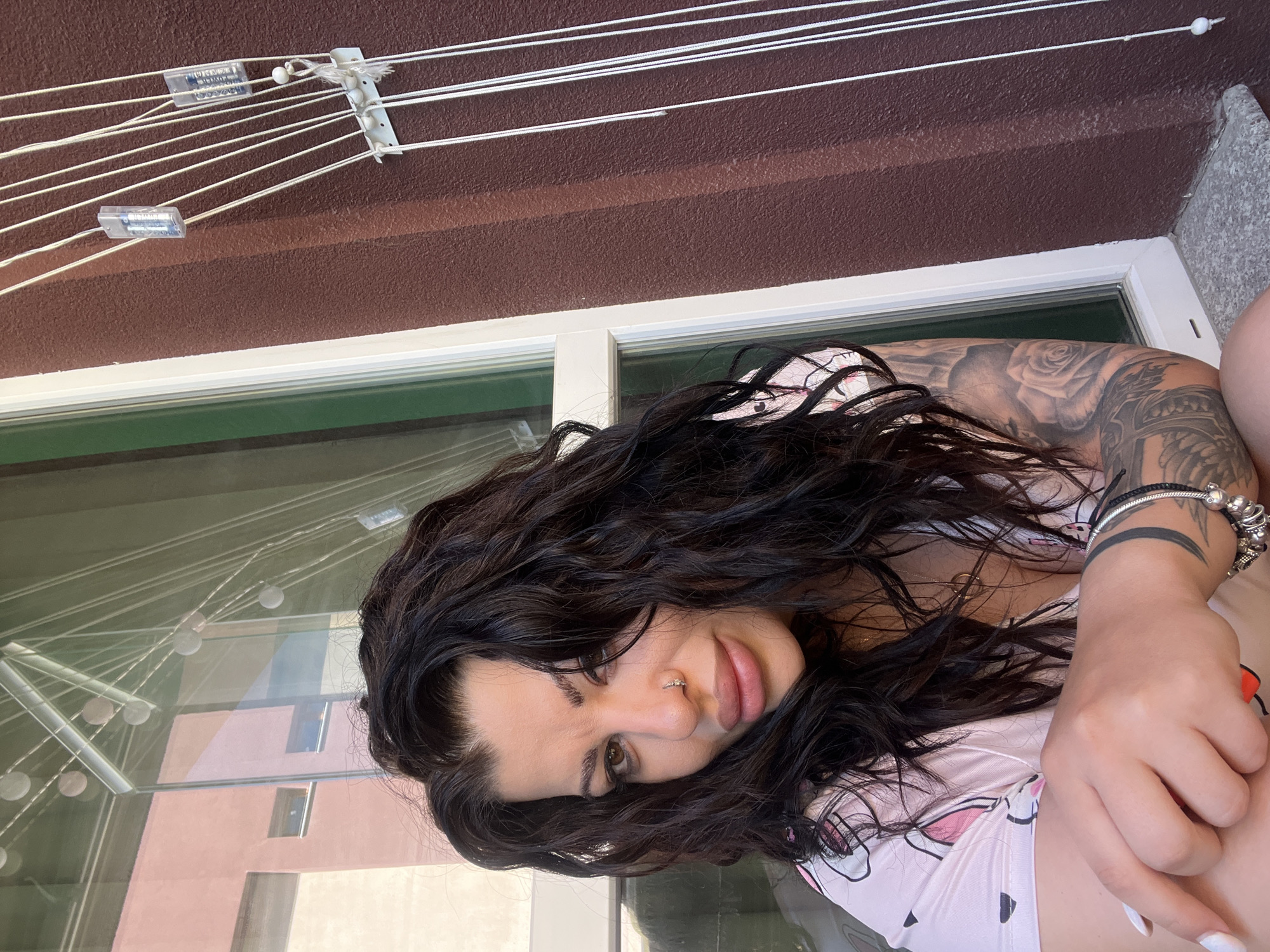 A young woman is pictured on a balcony in Plovdiv, Bulgaria. She has dark, curly hair, a nose piercing, and several tattoos on her right arm. She is wearing a pink patterned top and several bracelets on her right wrist. The balcony overlooks a street with a modern building reflected in the window. A drying rack with clotheslines is attached to the brown-textured wall above the window.