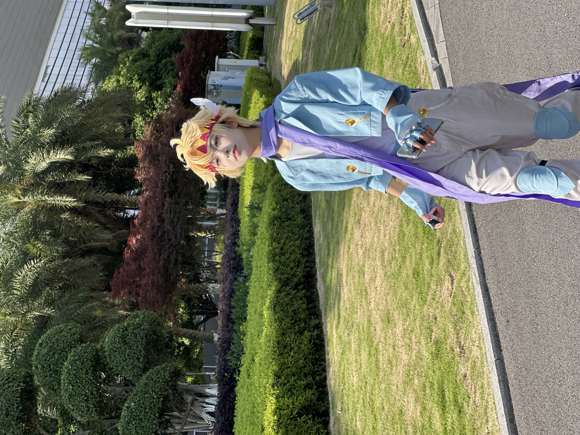 A person dressed in cosplay attire stands outdoors in Liangjiang, China. The cosplayer, with blonde hair, a patterned headband, and a blue jacket, is positioned on a patch of green grass beside a paved pathway. They are holding a smartphone in one hand and an object in the other. In the background, a variety of green trees and bushes, along with a building, are visible.