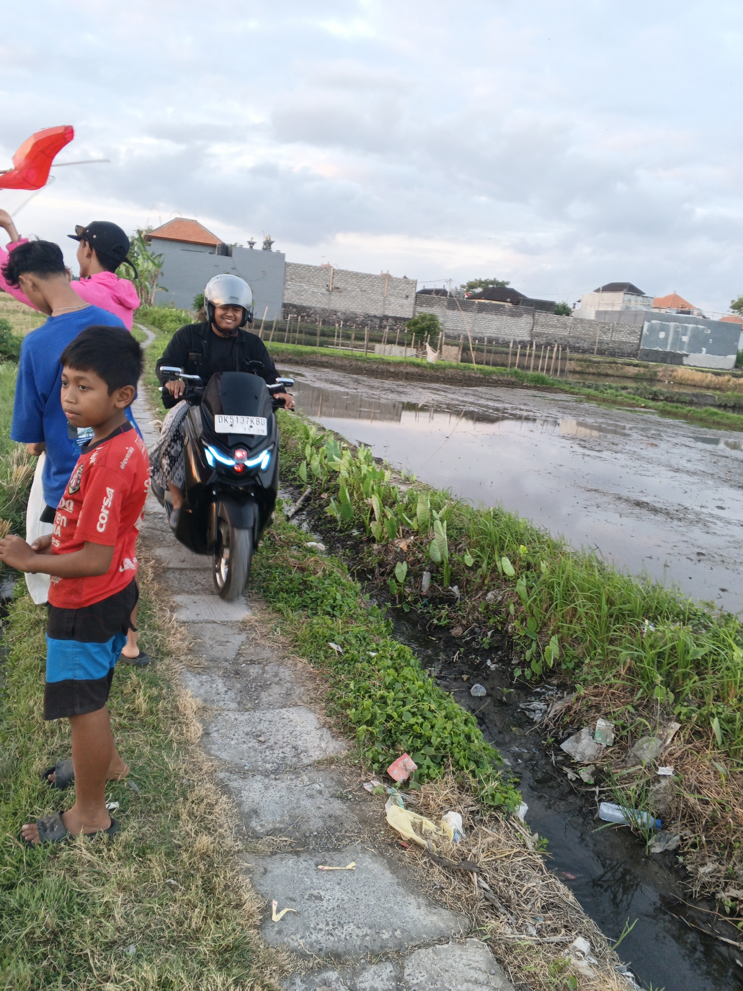 The image depicts a scene in Sukawati, Indonesia, likely a rural or semi-rural area with agricultural fields. In the foreground, a young boy wearing a red t-shirt and blue and black shorts stands on a grassy path. Behind him, two other individuals are partially visible. To the right of the path, a person wearing a helmet is riding a black motorcycle. The motorcycle has glowing blue headlights and a license plate reading "DK5137KBU". The path runs alongside a muddy field or waterlogged area, bordered by dense green vegetation and a small, debris-filled water channel. In the background, there are buildings constructed with brick and concrete walls, and some wooden fencing, suggesting ongoing construction or development. The sky is overcast with clouds. The overall atmosphere appears to be late afternoon or early evening, given the lighting conditions.