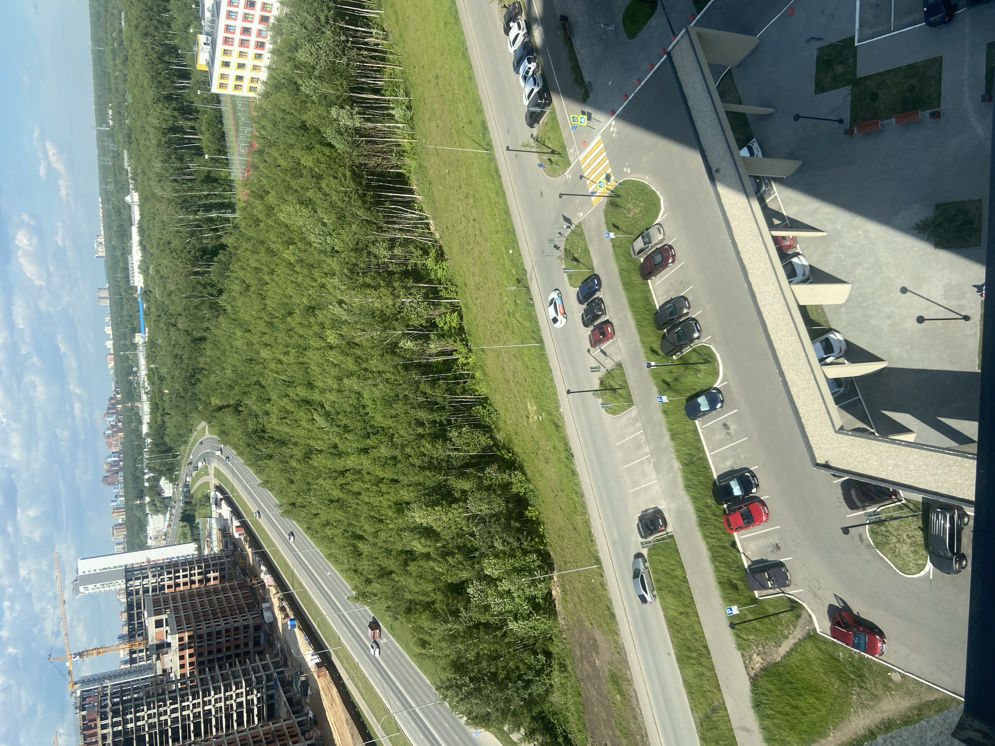 An aerial view captures a dynamic urban landscape, likely in Kazan, Russia. In the foreground, a multi-story building under construction features exposed concrete structures and scaffolding, with a prominent yellow crane dominating the sky to the left. Adjacent to the construction site, a multi-lane highway stretches into the distance, with several cars visible. A dense forest of green trees borders the highway, creating a stark contrast between the urban development and the natural environment. Further in the distance, a cluster of high-rise buildings can be seen, indicating a sprawling city. The sky is partly cloudy, with white clouds scattered across a blue expanse. The lower portion of the image shows a parking lot with several parked cars, including a red SUV, a black sedan, and a silver hatchback. A covered walkway with supporting pillars is also visible, casting a shadow on the pavement. A small patch of grass with a bench is situated nearby.