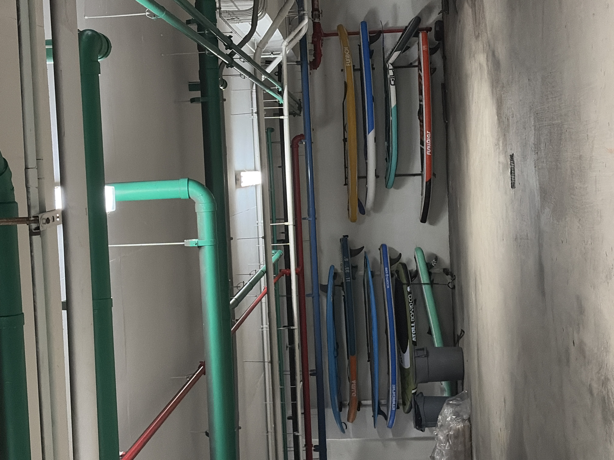 The image depicts an interior space, likely a storage or utility area, in a building. A prominent feature is the network of pipes running along the wall and ceiling. The pipes are colored green, white, and red. Several paddleboards are mounted on the wall, arranged vertically in racks. These paddleboards have varying colors and brands printed on them, including “FUNBOY” and “ATOLL BOARD CO”. The wall appears to be concrete and is mostly gray. There is also some refuse at the bottom of the paddleboards. Given the presence of paddleboards, a coastal setting might be implied, and the user has indicated Key Biscayne, United States of America, as the location. There are no people visible in the image.