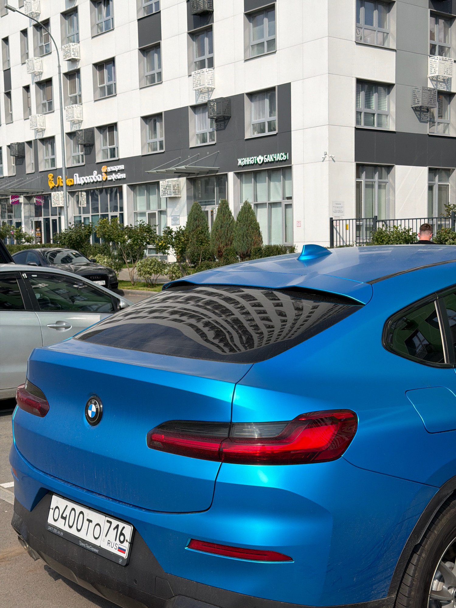 In this image, a blue BMW SUV is parked in front of a modern building with a glass facade and white and gray siding. The car's license plate is visible, reading "0400 TO 716". Another car, a dark sedan, is parked behind the BMW. The building appears to be a commercial or residential complex, with several shops on the ground floor, including one with a sign that reads "Женят бакчас" and another with Cyrillic text that translates to "Pirogovaya". The scene is set in an urban environment, possibly a city, with a streetlight and trees visible. The reflection of the building on the car's window suggests it's a sunny day.