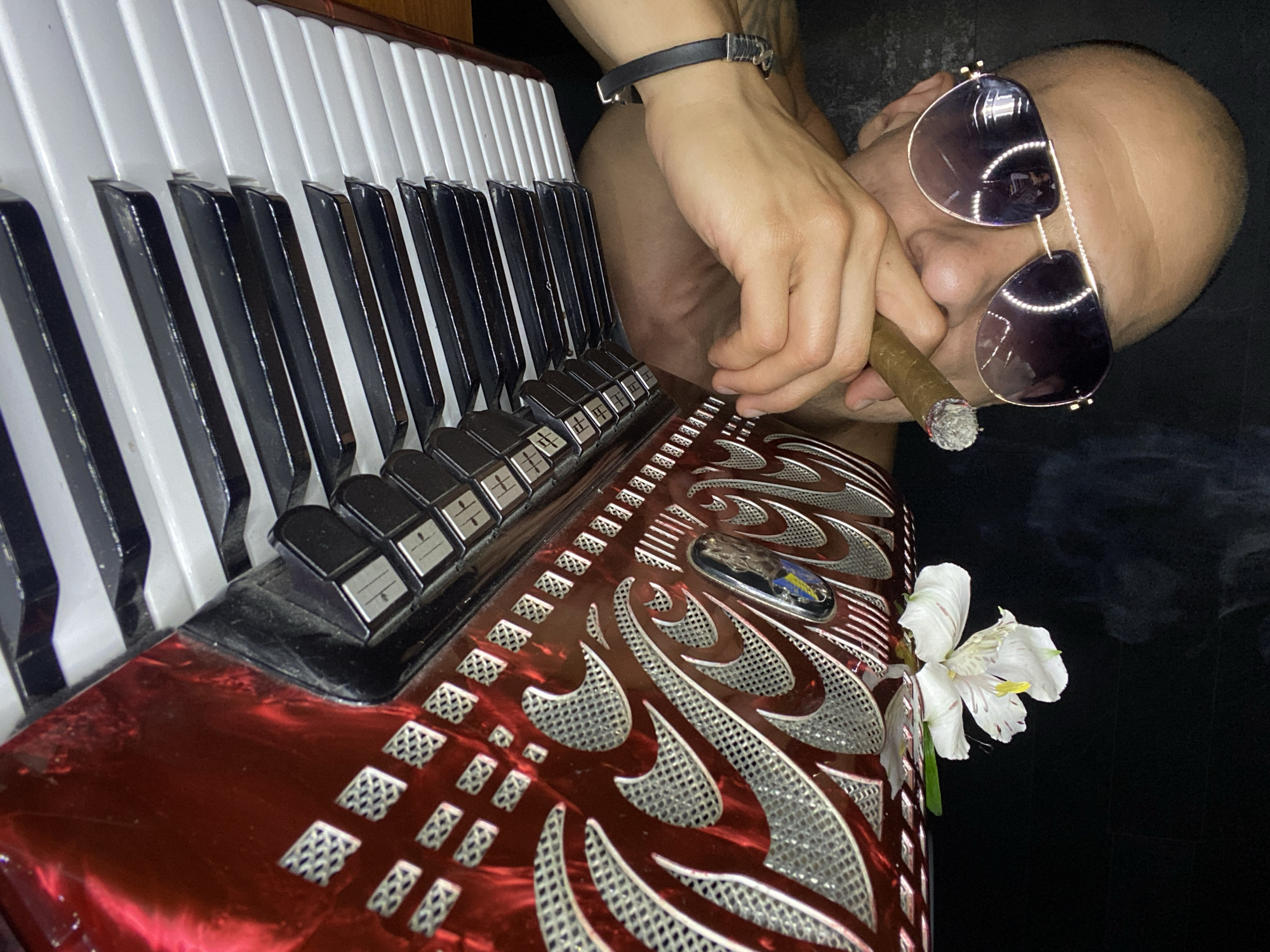 The image shows a close-up of a bald man wearing aviator sunglasses and holding a lit cigar. He is leaning towards a red accordion, with his right hand resting on its keys. A white flower is placed on top of the accordion. Smoke from the cigar is visible in the background. The scene appears to be indoors, with a dark background. There is no visible indication of the location being in Pleven, Bulgaria.