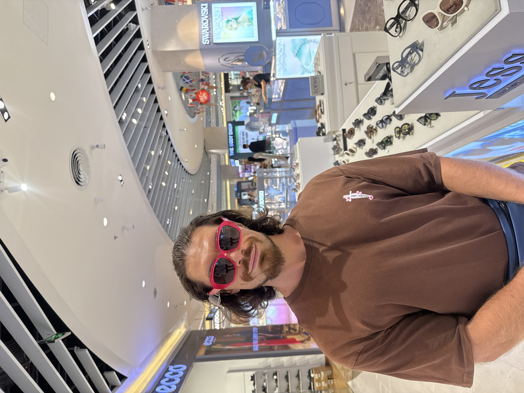 The image captures a man in a brown t-shirt and blue fanny pack wearing oversized pink sunglasses. He is standing in what appears to be a shopping mall, likely an eyewear store, as evidenced by the displays of glasses and the presence of multiple retail outlets in the background. One prominent sign reads "SWAROVSKI." The man is smiling and looking directly at the camera. The background shows a bright, modern interior with curved architecture and overhead lighting. The location is identified as Nha Trang Ward, Vietnam.