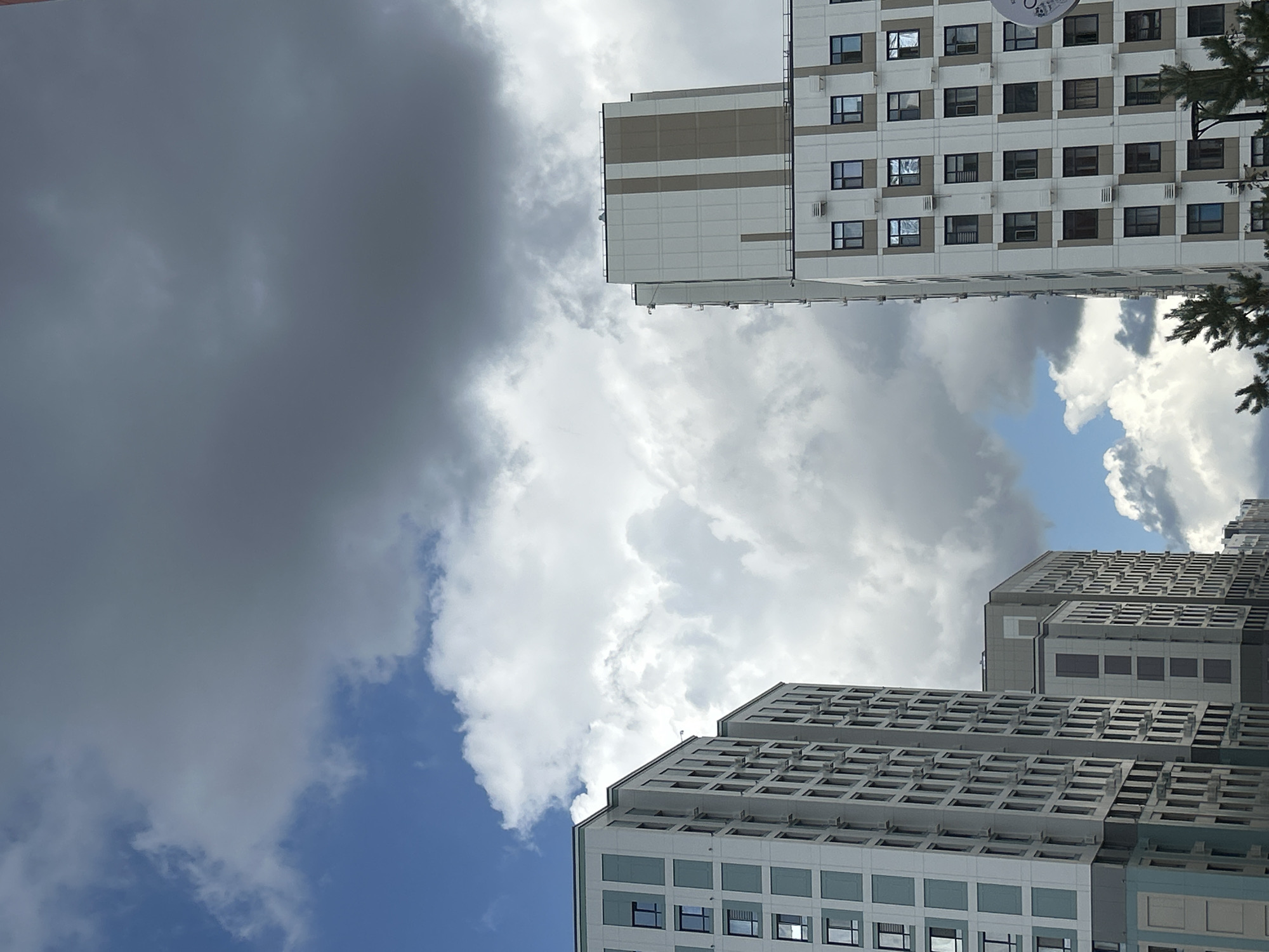 The image shows the exterior of modern apartment buildings against a sky filled with both bright white and dark gray clouds. The buildings are tall and appear to be constructed with light-colored facades, featuring numerous windows. Some buildings have vertical accents of a darker color. The overall scene suggests an urban environment, likely in Astana, Kazakhstan, given the architectural style and the prevalence of such high-rise residential complexes in the city. There are no people visible in the frame. The lighting indicates it might be daytime, with the contrast between the sunlit clouds and the shadowed ones suggesting a dynamic weather condition.