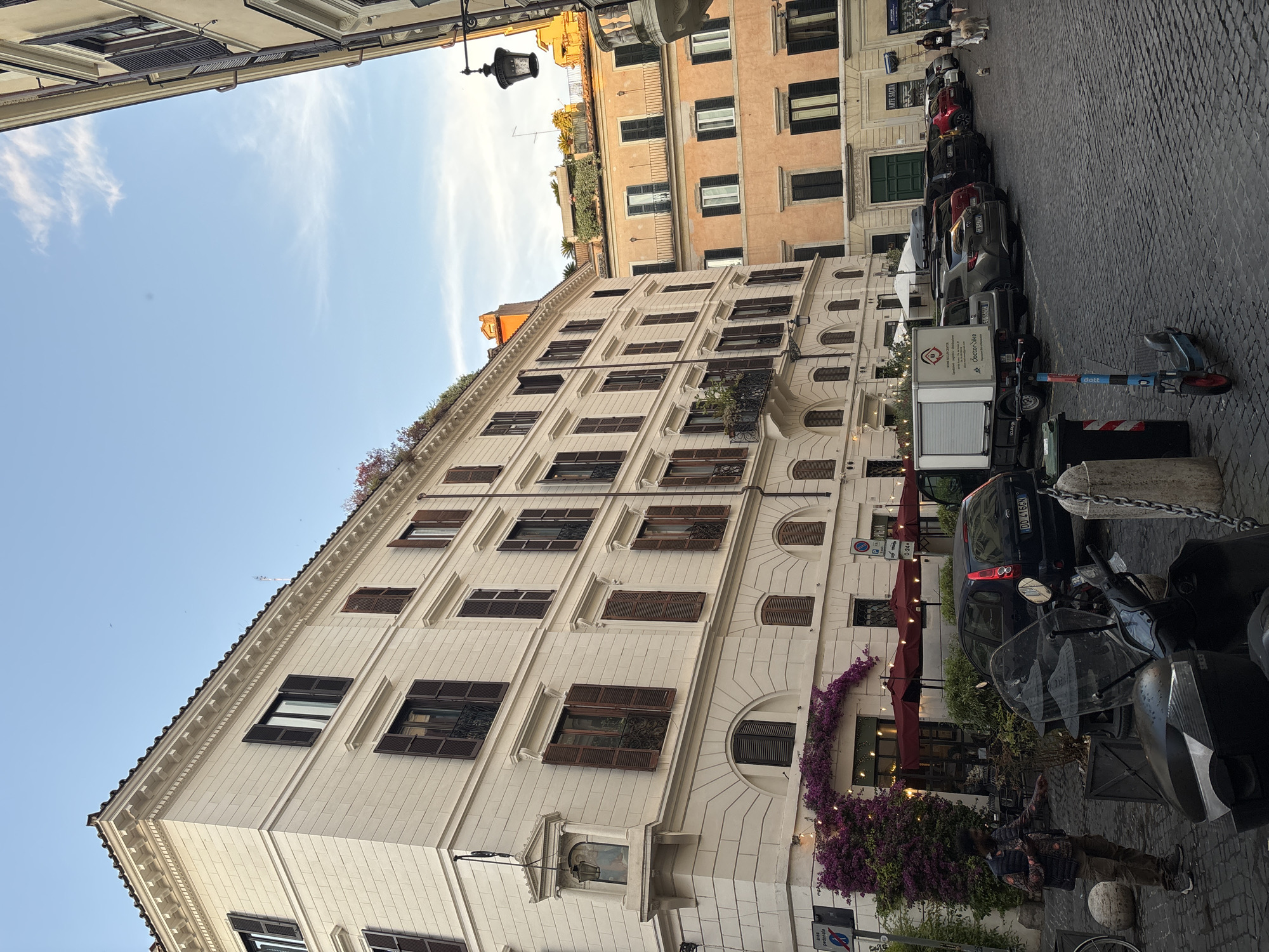 The image shows a street scene in Rome, Italy. A tall, multi-story building with light-colored facade and numerous windows with dark shutters dominates the left side of the frame. The building has architectural details such as decorative moldings and arched windows on the lower levels. A cascading bougainvillea with purple flowers adorns the building near ground level.
Parked cars and scooters line the right side of the street. A few people are visible near the building, including a person standing close to the bougainvillea. The street is paved with cobblestones. The sky is partly cloudy and blue.