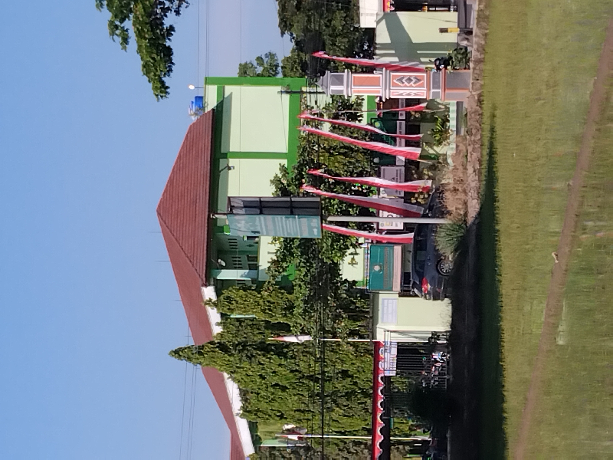 The image shows the exterior of a two-story building, likely a school or government facility, painted light green with a reddish-brown tiled roof. It is located in Papahan, Indonesia. In front of the building, there is a decorative pillar with alternating grey and red-orange bands. Several red and white Indonesian flags are displayed along a fence line. A dark-colored SUV is parked to the right of the entrance. Tall evergreen trees are planted along the building's facade. In the foreground, a partially flooded rice field is visible under a clear blue sky, with a dirt path running through it. The scene appears to be captured during the daytime with ample sunlight.