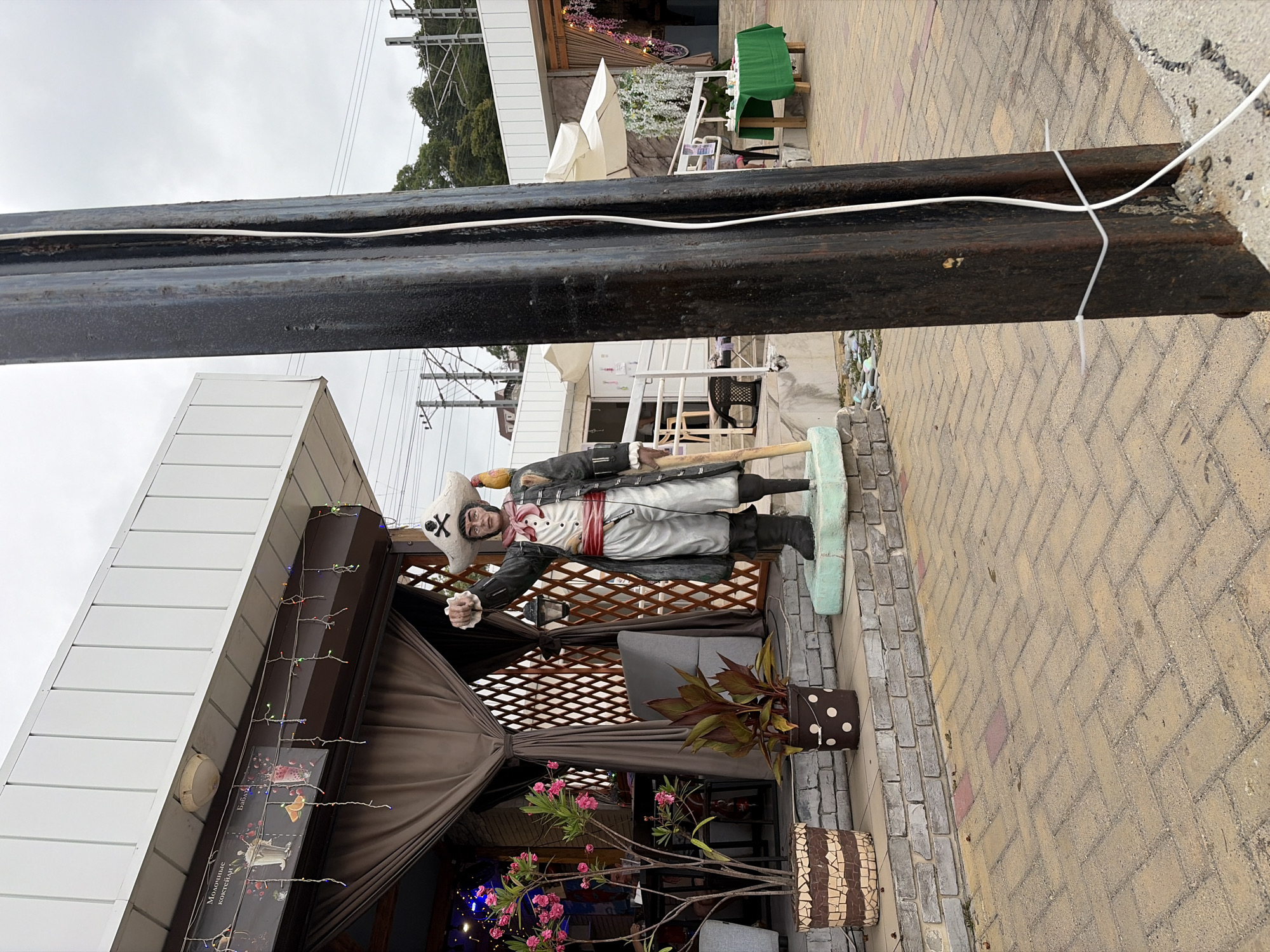 A statue of a pirate, holding a lantern and a staff, stands on a pedestal in front of an outdoor cafe. The pirate wears a tricorn hat, a ruffled shirt, a sash, and a long coat. A parrot is perched on his shoulder. Pink flowers bloom in pots nearby. The cafe has tables and chairs set up outside, some covered by umbrellas. A green tablecloth covers a table in the background. The scene is set on a paved walkway. The overall setting suggests a tourist area in Sochi, Russia, possibly near the coast, given the presence of an outdoor dining area and what appears to be train tracks in the background. A large metal pole, likely a utility pole or part of a structure, is in the foreground.