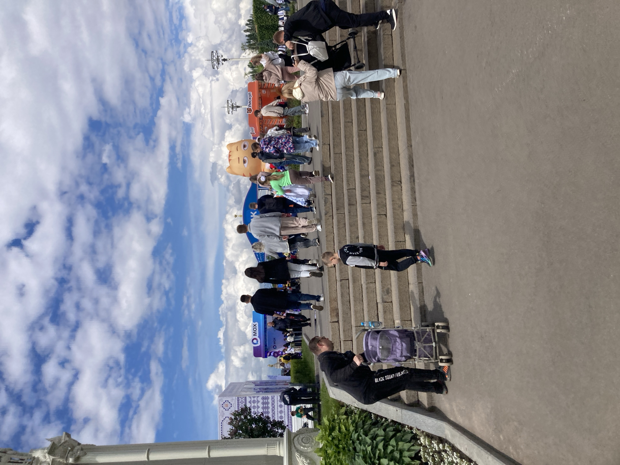 This image captures a vibrant scene on a sunny day in Moscow, Russia, likely at VDNKh park due to the distinctive architecture and the presence of a large crowd. People of varying ages are moving down a wide set of steps. On the left, there are promotional materials and a large cartoon cat figure, potentially indicating an event or festival. Some people are pushing strollers. A man wearing black clothing and "BLACK SQUAD PROJECTS" is also pushing a stroller. The architecture on the far left suggests a historical building. A bright blue sky dotted with fluffy white clouds provides a backdrop to the scene.