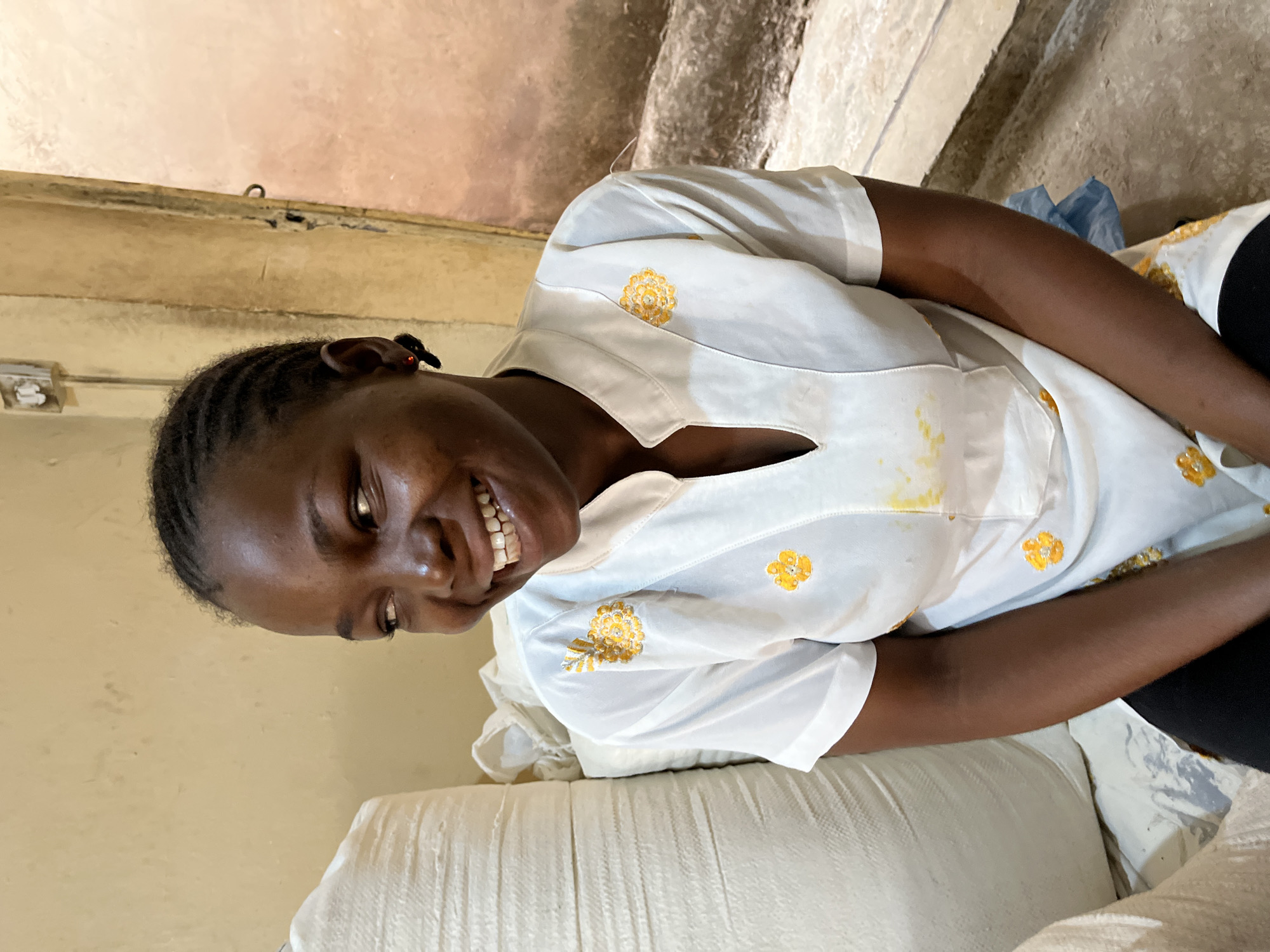 A young Black woman with braided hair is smiling and sitting indoors in Jos, Nigeria. She is wearing a white shirt with small, yellow floral embroidery and a notched neckline. She is sitting on what appears to be a large, white, fabric-covered object, possibly a sack or cushion. The background includes a wall painted beige, a light fixture, and a concrete structure. The light is diffused and indirect.