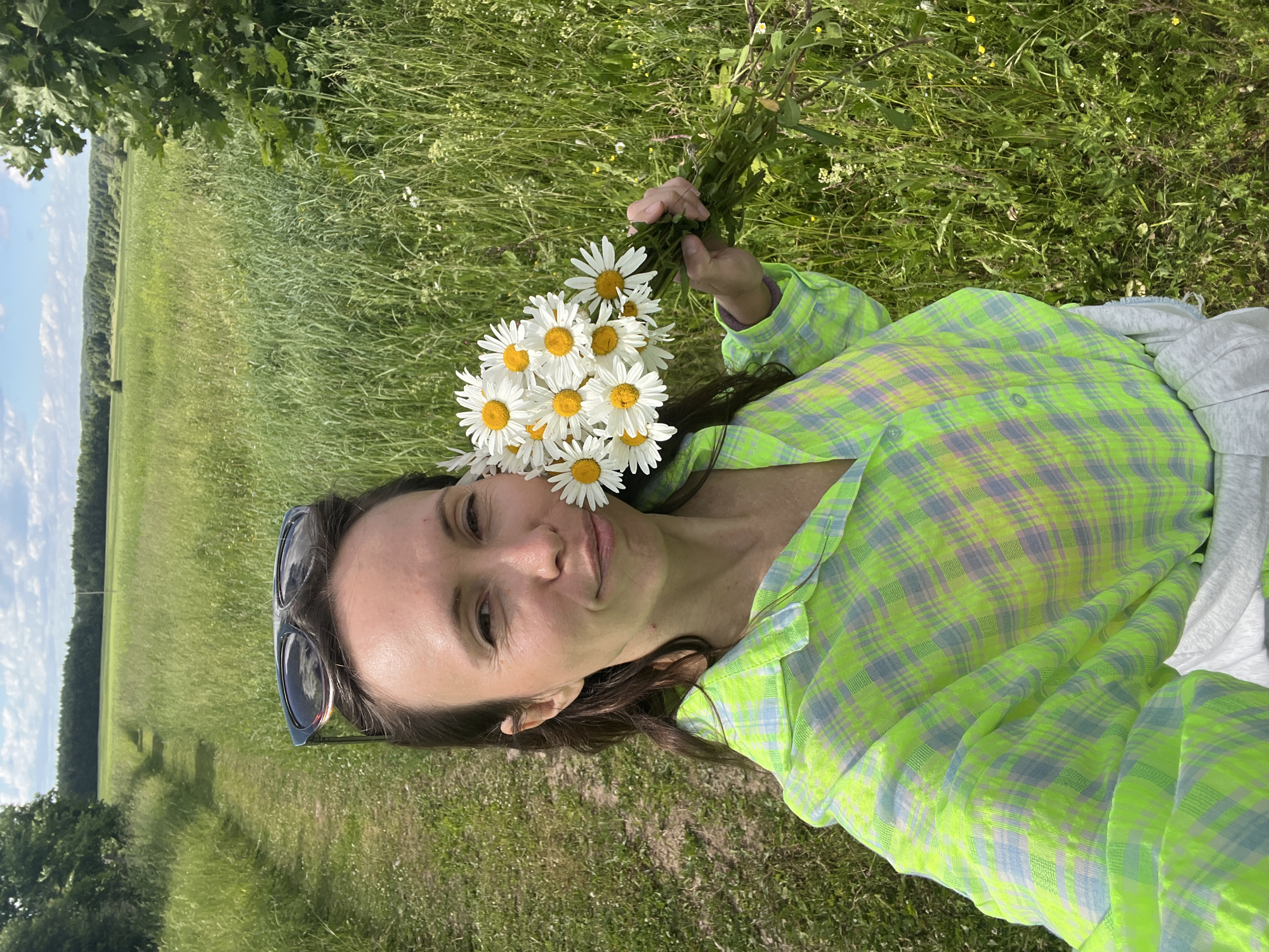 In this selfie taken outdoors, a woman with brown hair pulled back and sunglasses perched on her head is holding a bouquet of daisies. She is wearing a neon green and gray plaid shirt. The background features a narrow dirt path leading through a tall, green grassy field, with trees and a distant tree line under a cloudy blue sky. The scene appears to be in a rural area, potentially near Пигозино, Russia, given the expansive natural landscape.