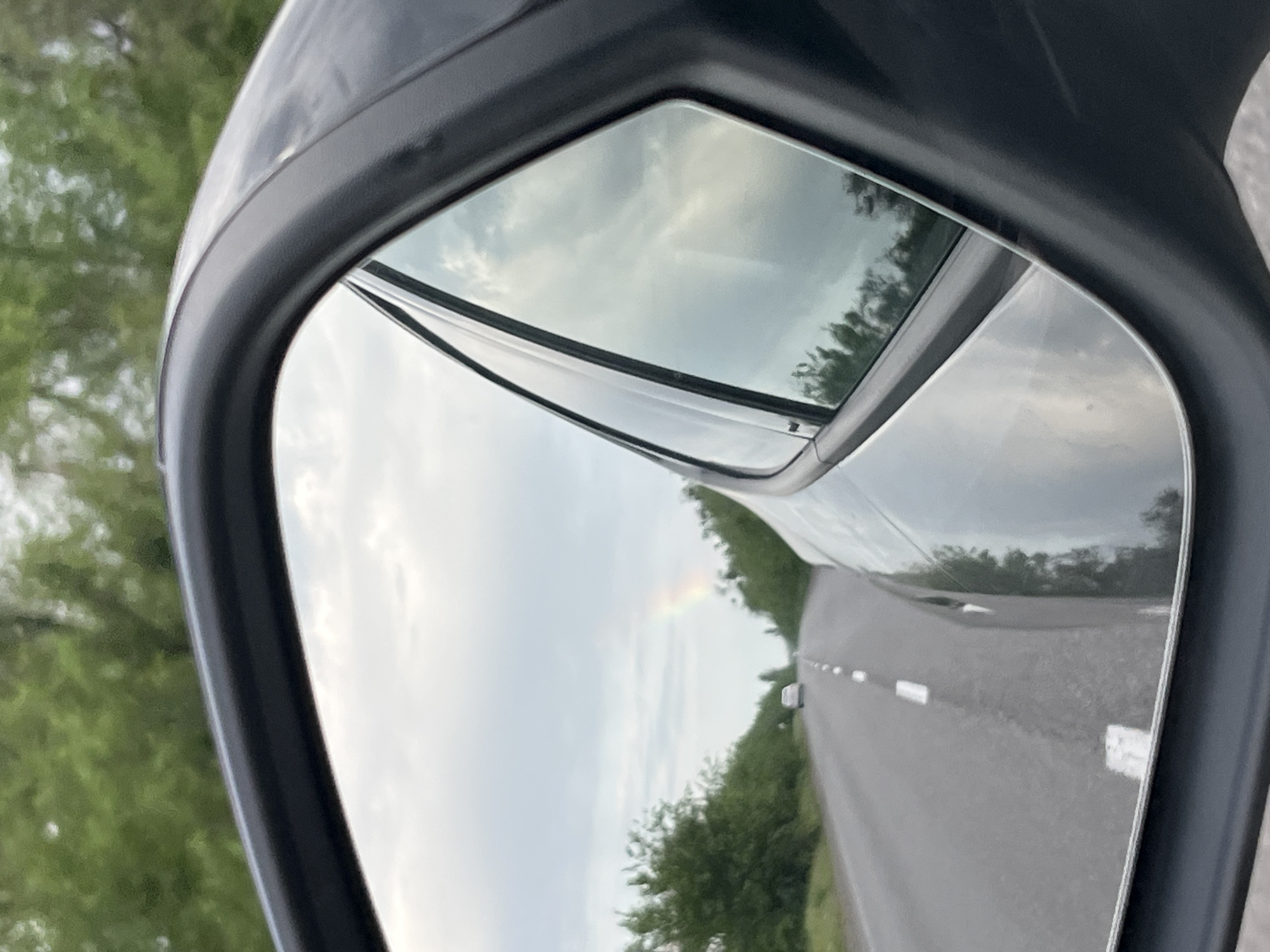 The image shows the reflection in a car's side mirror. The reflection includes a road with white lane markings, green vegetation on either side, a car in the distance, and a cloudy sky with what appears to be a rainbow. The location is unknown and not determinable from the image. There is no indication of Sofiivka, Ukraine.
