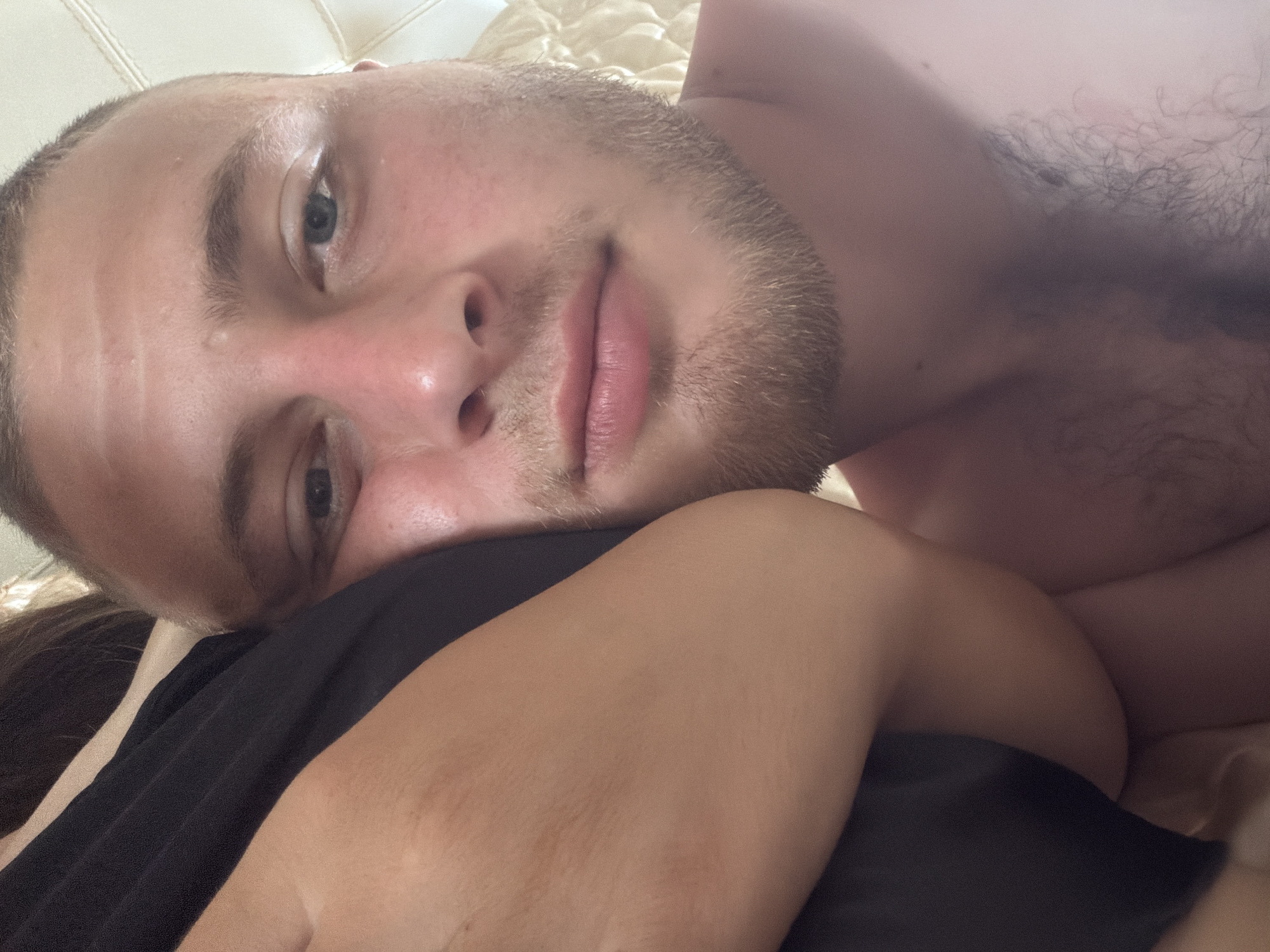 The image shows a close-up, personal view of a young man with short hair and stubble, resting his head on another person's shoulder, which is covered by a black garment. The man's eye is visible, looking towards the camera. The background appears to be a softly lit, textured surface, possibly bedding or upholstery. The overall impression is intimate and relaxed. The location in Strășeni, Moldova is not discernible from the visual information provided.
