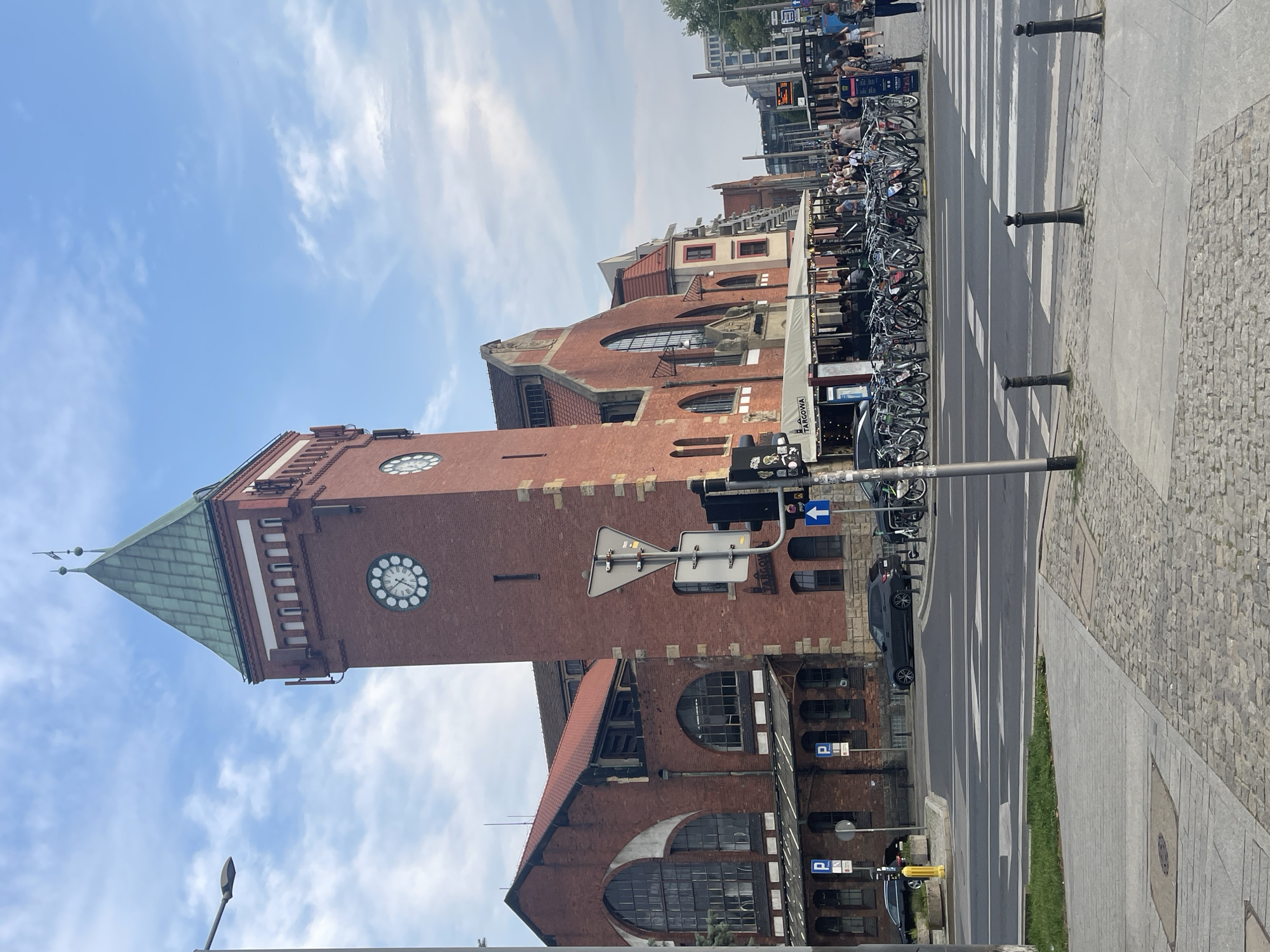 The image shows a street scene in Wrocław, Poland, with the historic Hall of the Merchants (Hala Targowa) building as the focal point. The building is constructed of red brick and features a prominent clock tower with a green roof. In front of the building, there is a street with a crosswalk, several cars, and a large number of bicycles parked along the sidewalk. People are visible walking and standing on the sidewalk. The sky is partly cloudy. The location appears to be a busy urban area with a mix of historical and modern architecture.