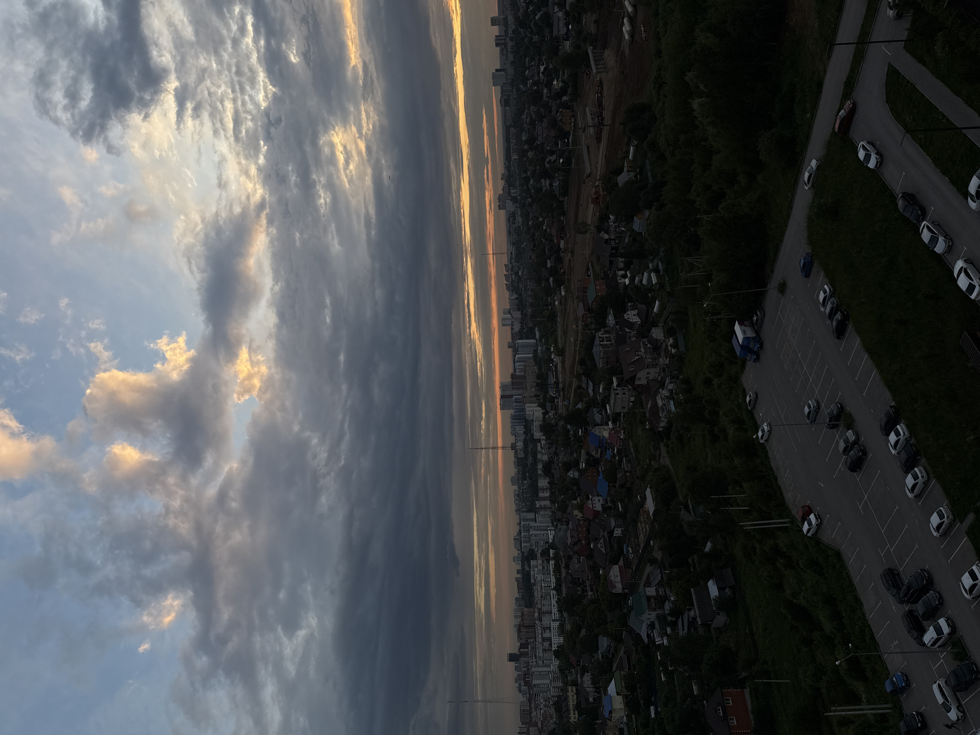 The image captures an aerial view of a city during a cloudy sunset, likely in Perm, Russia, given the architectural style of the buildings and the presence of two tall radio towers that are characteristic of Soviet-era urban planning. The sky is dominated by a mix of dark gray and lighter, wispy clouds tinged with orange and yellow hues from the setting sun. Below the sky, a sprawling cityscape unfolds, featuring a dense collection of residential buildings, ranging from high-rise apartment blocks to smaller, detached houses with pitched roofs. The city is interspersed with patches of green trees and vegetation, indicating a mix of urban and suburban areas. In the foreground, a large, paved parking lot is visible, populated with numerous parked cars of various colors and models. The overall scene is one of a typical urban landscape at dusk, with the natural beauty of the sunset contrasting with the man-made environment.