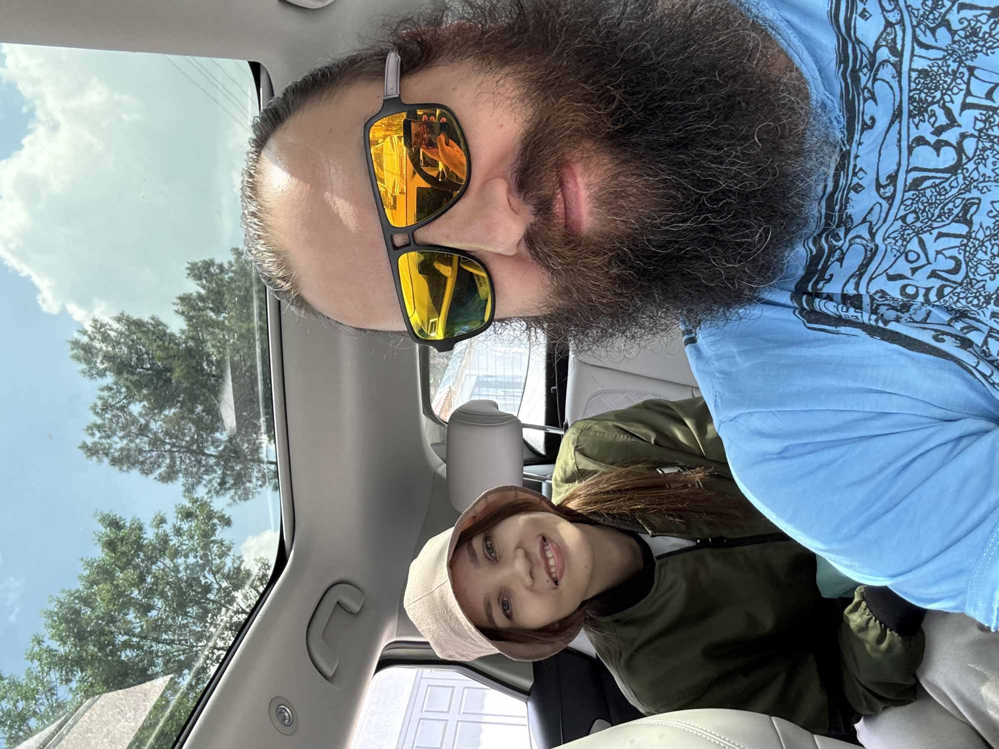 Two individuals are inside a car. The person in the foreground, closer to the camera, is a bearded male wearing sunglasses and a blue patterned shirt. The reflection in the sunglasses shows a hand holding a phone. The second person, possibly female, is wearing a bucket hat and an olive green jacket. The environment visible through the car windows shows greenery and buildings. It is difficult to ascertain the exact location in Tashkent, Uzbekistan.