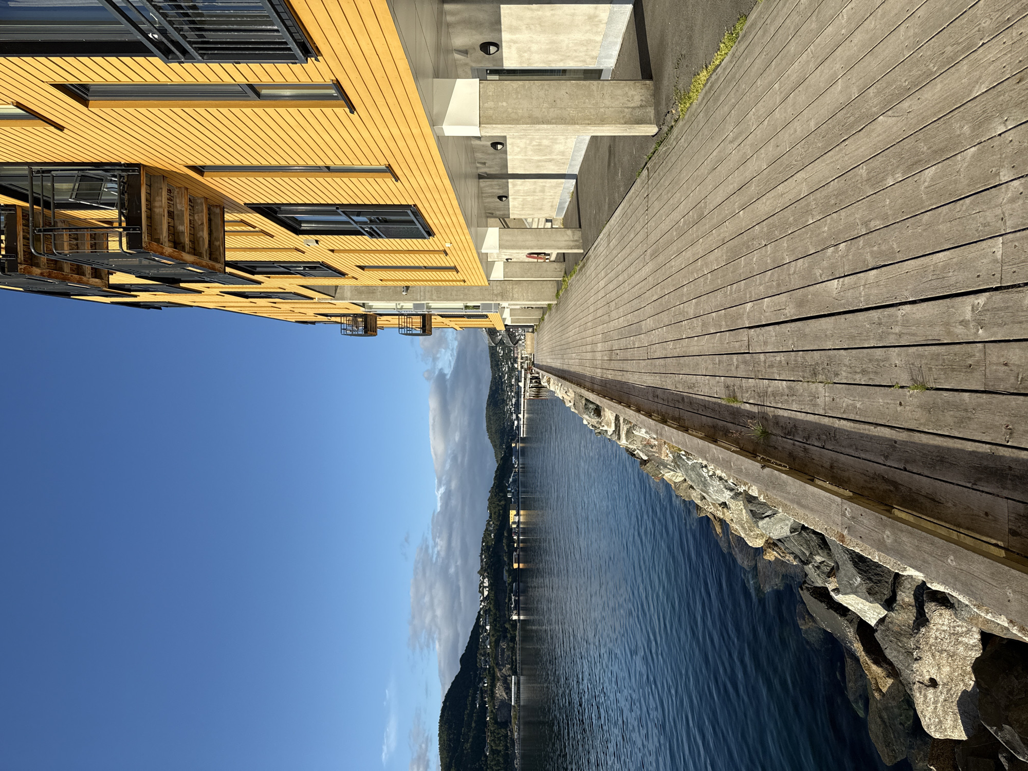 A waterfront promenade in Harstad, Norway, runs alongside a bright yellow building with balconies. The promenade is constructed of weathered wooden planks and is bordered by a rocky embankment. The water, a deep blue, gently laps against the rocks. In the background, a hillside town with houses and buildings is visible under a clear blue sky with a few scattered clouds. The scene is devoid of any people.