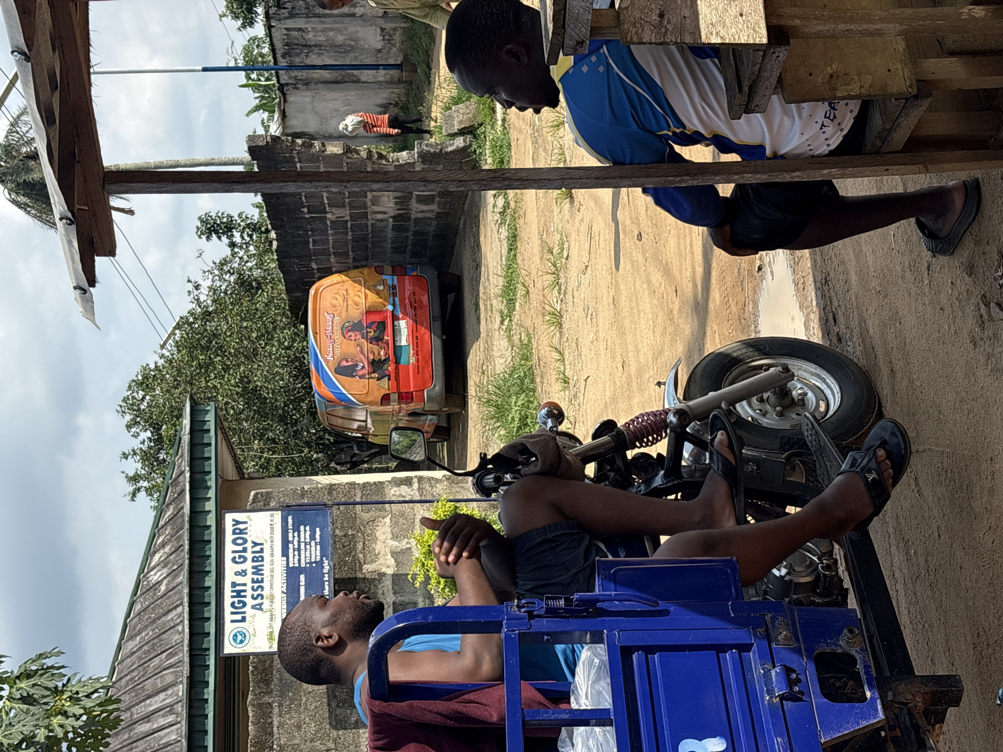 The image depicts a street scene in Ikot Ekpene, Nigeria. A man is sitting on a blue motorized tricycle. Beside him is a sign for "Light & Glory Assembly," a Christian church located on Kotanak Off Abanpo Road by Christian Sec. Sol Abiakpo Kot Esser. Behind the tricycle is a colorful van with an advertisement on it. A second man is sitting on a wooden bench. A person is lying on a concrete ledge in the background.