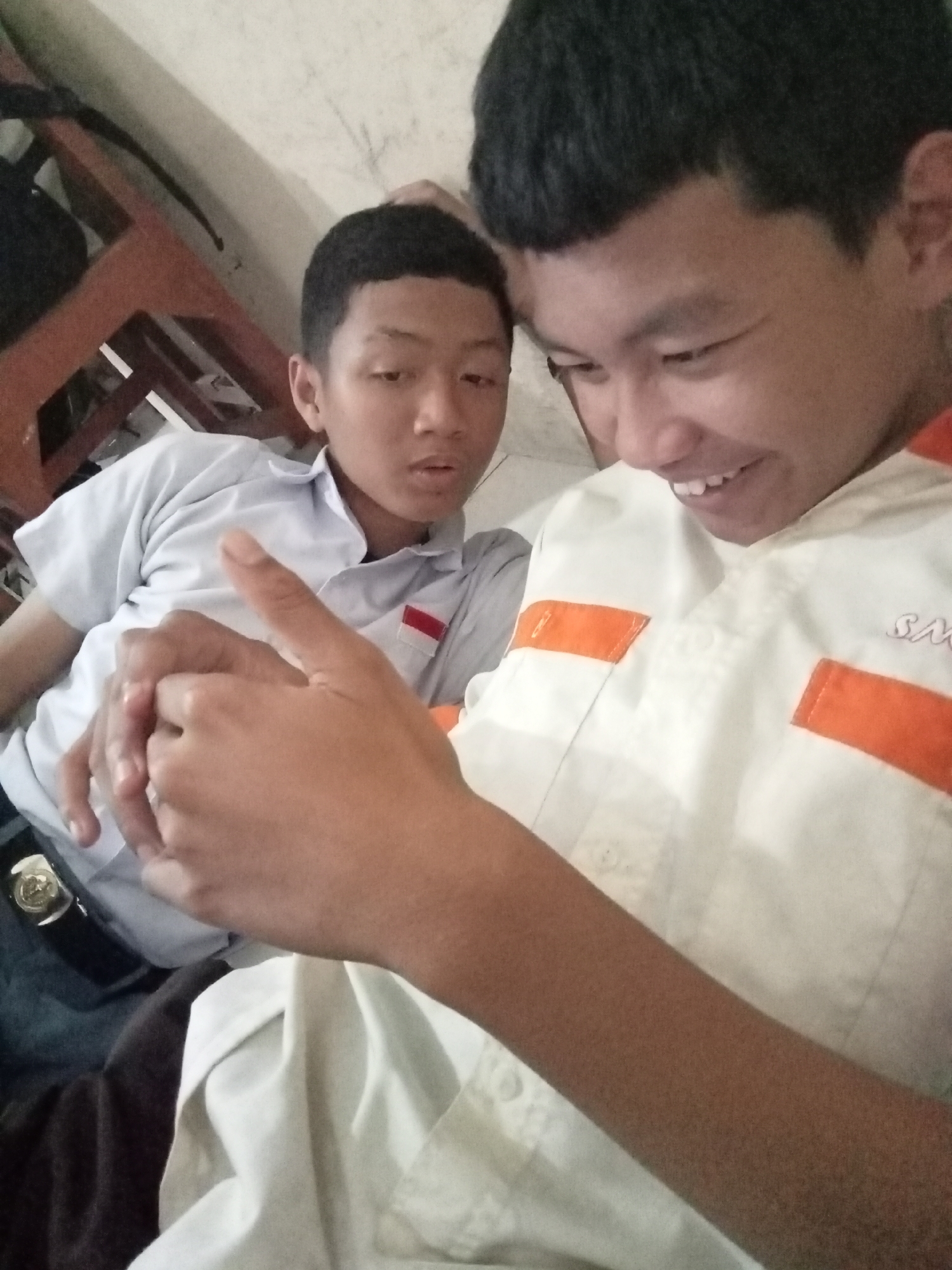 Two young males, possibly teenagers, are shown in a close-up selfie. The male in the foreground is smiling and looking towards the camera, while the male in the background is looking slightly to the side. Both are wearing uniforms. The male in the foreground has a white shirt with orange accents and some visible text on the chest. The male in the background is wearing a light blue shirt with a red and white insignia on the chest. Their hands are visible in the foreground, with one appearing to be pointing. The background is indistinct but suggests an indoor setting with what appears to be furniture. The lighting is dim, and the image quality is not high. There is no discernible information about the location being Secang, Indonesia.