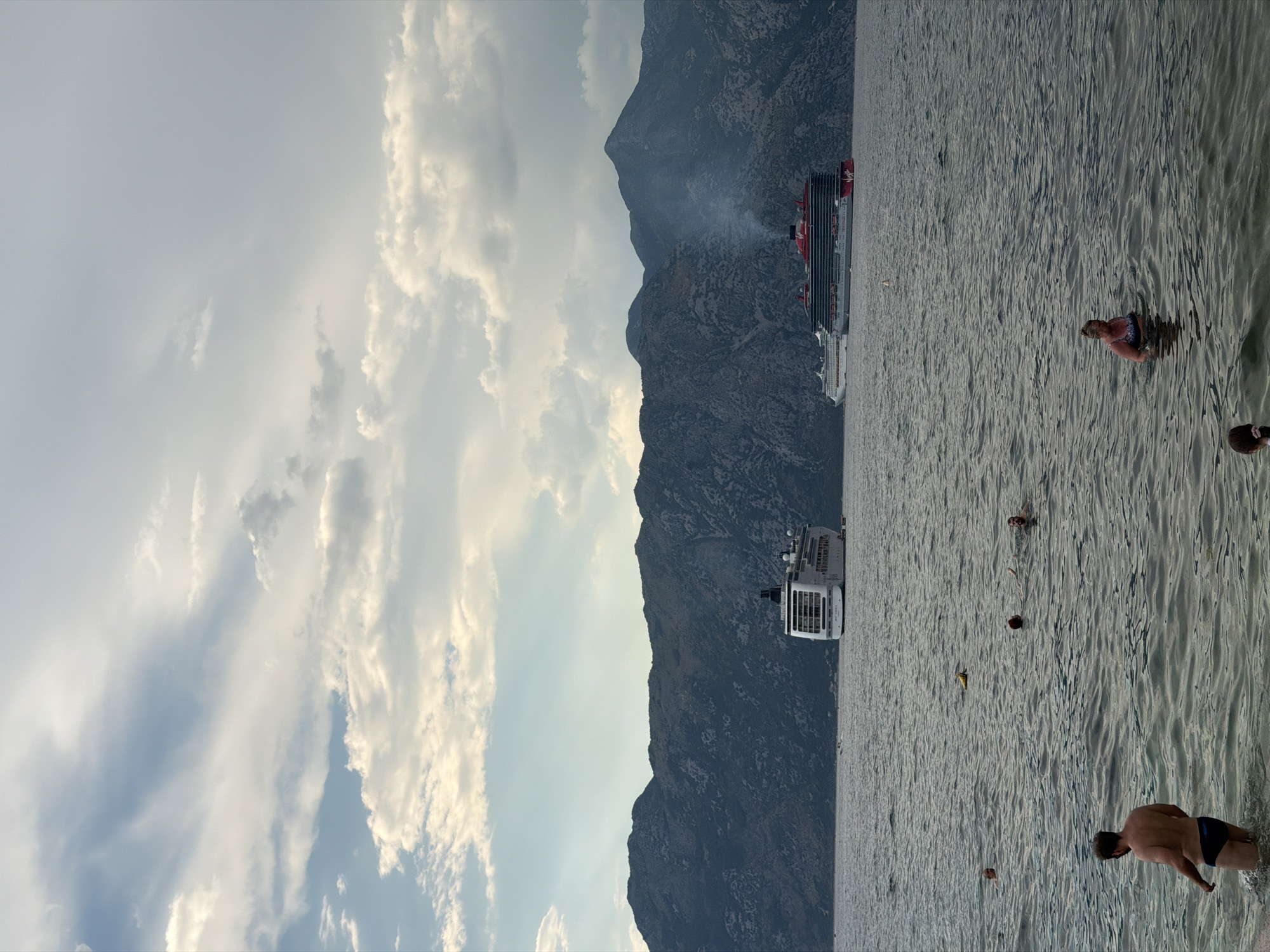 This is a photograph of the Bay of Kotor, taken from the shore in Dobrota, Montenegro. The image depicts a body of water with several people swimming or standing in it. In the background, a range of dark, mountainous terrain rises steeply from the water. Two large cruise ships are visible on the water, one closer to the shore and the other further out. The sky above is partly cloudy, with some shafts of sunlight breaking through. The overall atmosphere is one of a relaxed, scenic vacation setting.