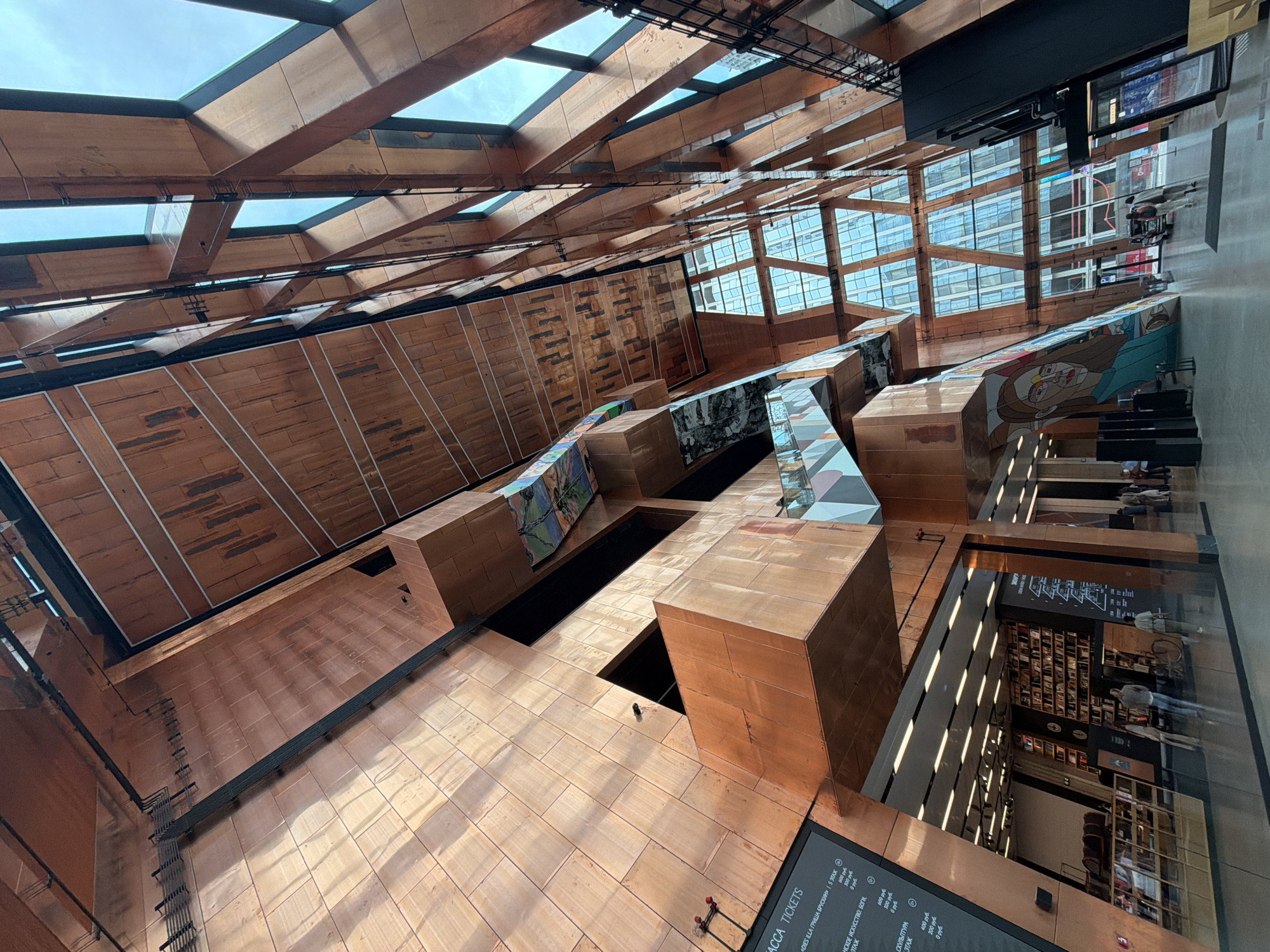This is a low-angle, interior shot of the GES-2 House of Culture in Moscow, Russia. The architecture features extensive use of copper-toned metal paneling and warm wooden elements, creating a striking, multifaceted interior space. The copper paneling is used on the walls and ceilings, often in large, angular sections. The wooden elements are seen in the ceiling structures and some internal walls, forming geometric patterns. Large glass windows form a significant part of the exterior wall, revealing a cloudy sky and the reflection of buildings on the opposite side.
Multiple levels and walkways are visible, suspended within the vast atrium. Some of these levels are clad in the same copper material, while others feature contrasting colors and materials, including patterned wallpaper or murals, and geometric designs on the railings. Escalators and staircases are integrated into the design, facilitating movement between levels.
On the ground floor, a large open space is visible. To the left, a section of the wall is covered with a large mural depicting a stylized portrait. Further back, a well-stocked bookshelf is visible, indicating a library or reading area. People are present throughout the space, walking around, some standing and observing. A ticket information board is mounted on a wall to the left, with Cyrillic text indicating various ticket prices and museum information. The overall impression is one of a modern, artistic, and spacious cultural center.