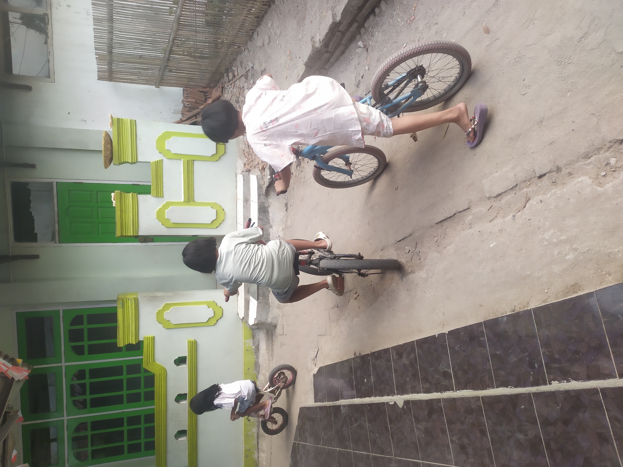 In Kragilan, Indonesia, three children are riding bicycles on a dirt path in front of a light green house with green trim. The house has large green windows and a green door. The child on the left is on a small pink bicycle, facing away from the camera. The middle child is on a darker bicycle, also facing away. The child on the right is on a blue bicycle, facing away from the camera, and is wearing a pink and white patterned shirt and shorts. The ground is dusty and uneven, with some tiled flooring visible on the left side of the image. A bamboo fence is visible on the right side of the house.