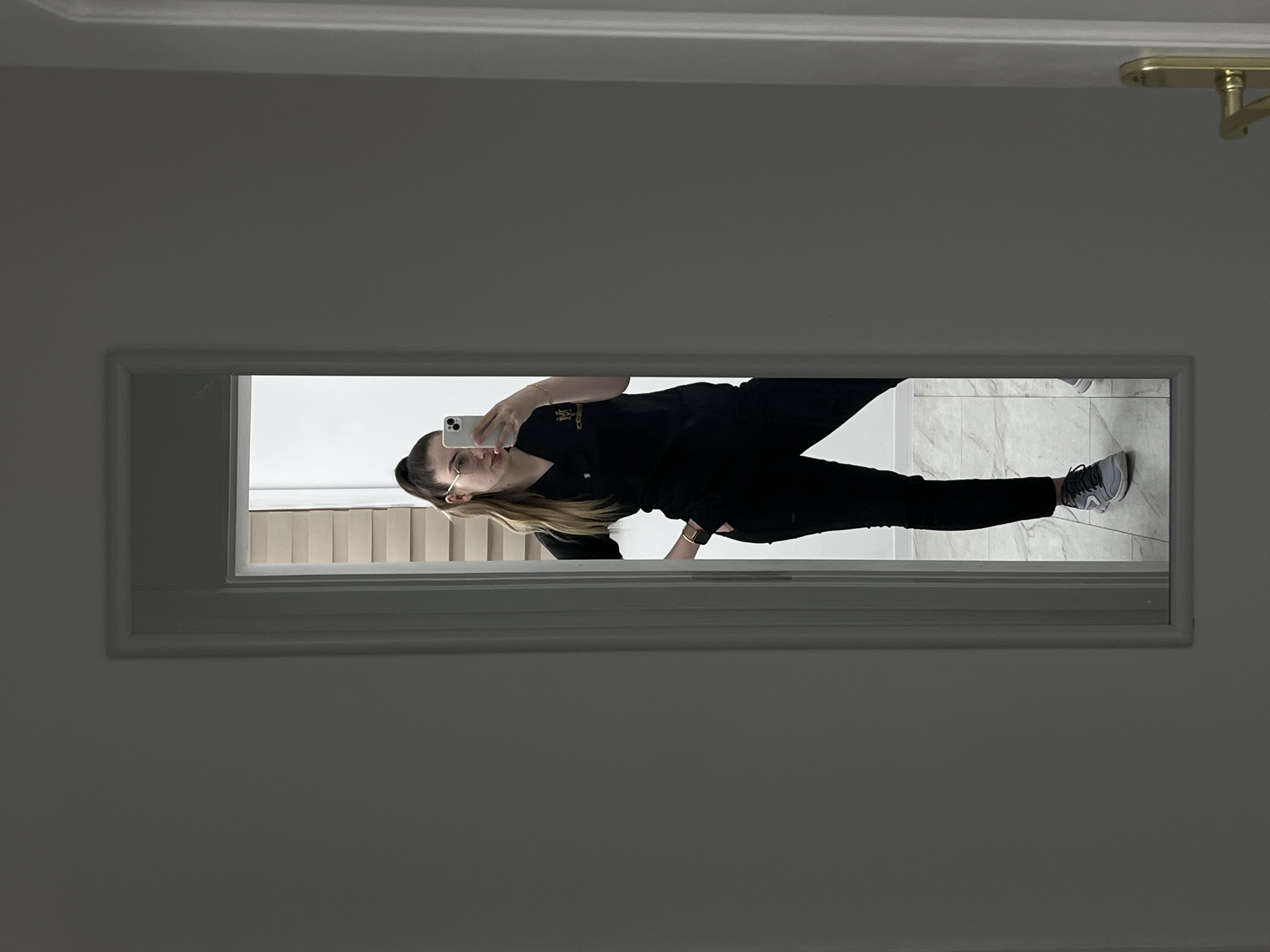 A woman, wearing black scrub top and pants with light blue athletic shoes, stands in front of a full-length mirror in an indoor setting. She is holding a white smartphone in her right hand, taking a selfie. Her hair is tied back in a ponytail, and she is wearing glasses. The mirror reflects a tiled floor and blinds on a window. The background appears to be a room with plain walls. The overall lighting is dim.