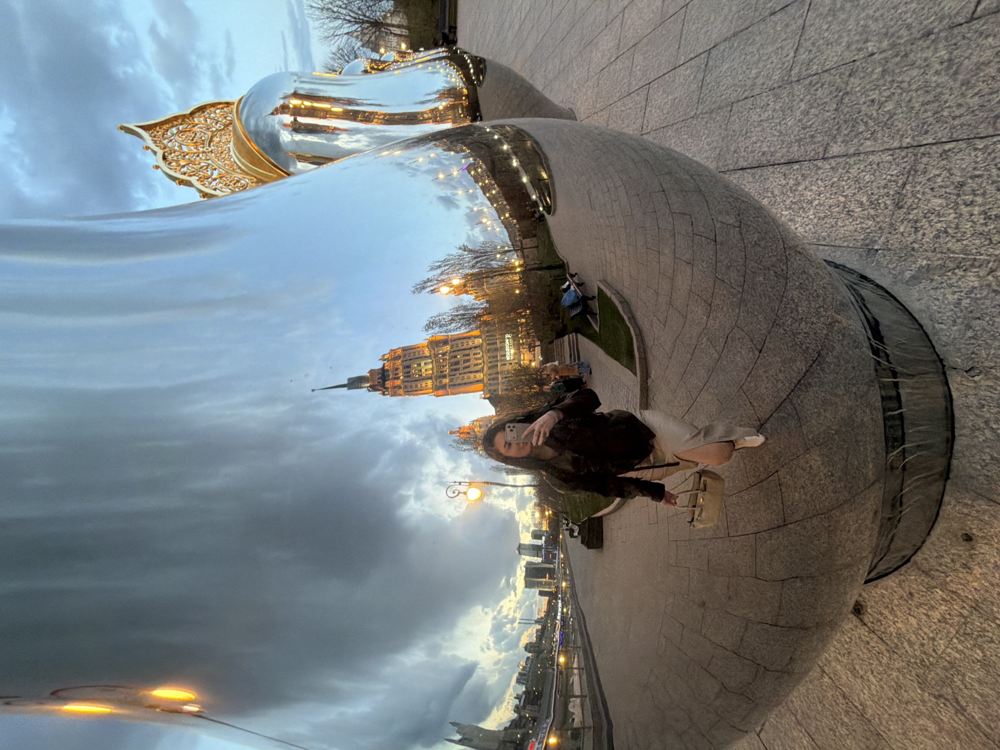 A woman stands in front of a silver sculpture, taking a selfie with her cell phone. The sculpture reflects a distorted view of the cityscape of Moscow, Russia, including a Stalinist skyscraper. The surrounding area appears to be a park or plaza with paved walkways. The sky is overcast.