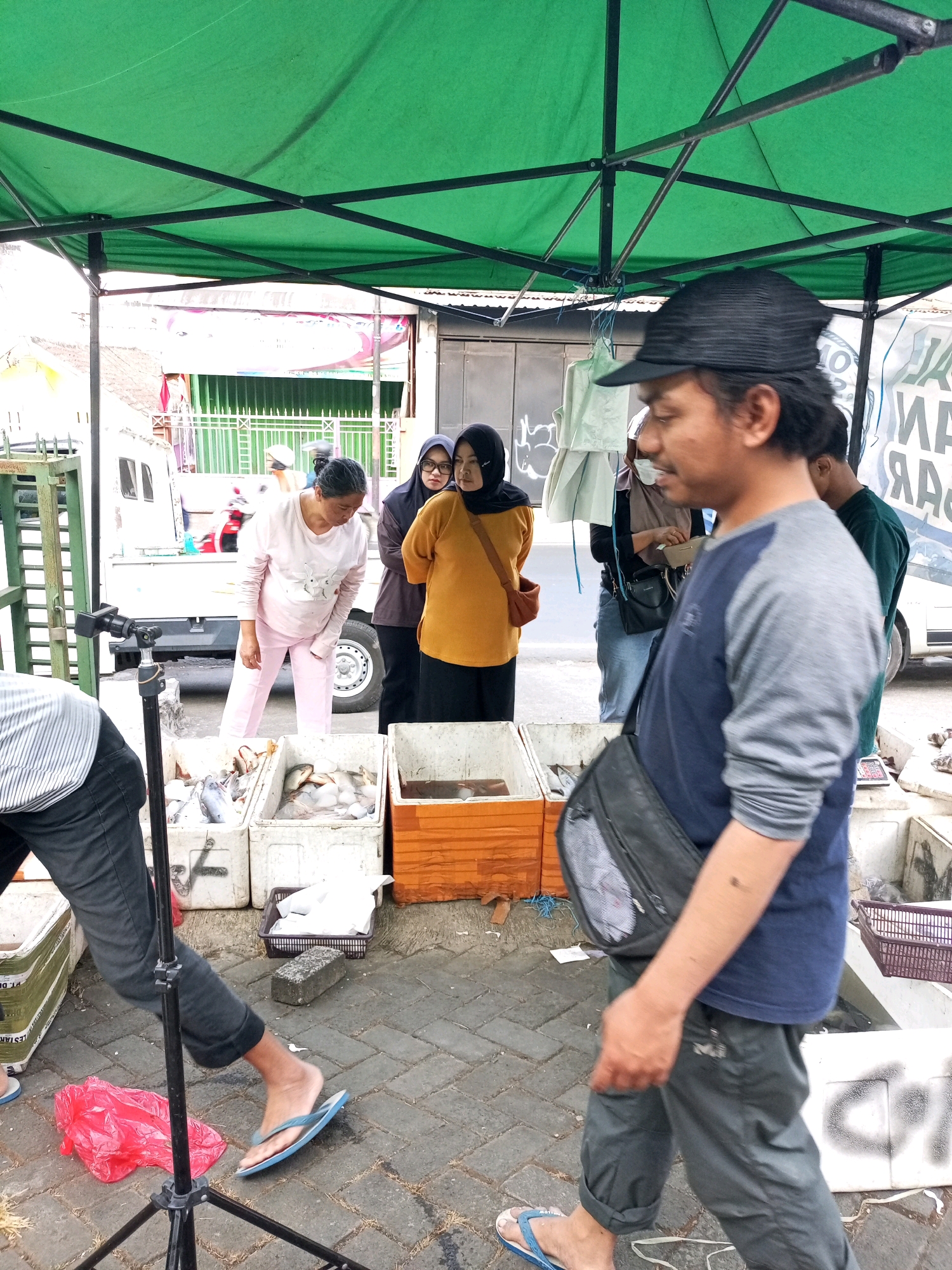 This image captures a street scene, likely a marketplace, under a green tarp in Batu, Indonesia. In the foreground, a man wearing a black cap, a blue and gray long-sleeved shirt, and dark pants with a black sling bag across his chest is walking towards the right. To his left, a person wearing a striped shirt and dark pants is partially visible, with a camera tripod in front of them.
Behind the man, several people are gathered. A woman in a pink shirt and pants is looking down at a crate of fish. Next to her, another woman in a dark hijab and a yellow top is standing, and beside her, a woman in a dark hijab and glasses is looking forward. Other individuals are visible further back.
The environment includes crates of fish laid out on the ground, a white truck, and various market structures. The ground is paved with brick-like stones. A red plastic bag and a gray brick are also present on the ground. The overall impression is that of a lively market setting.