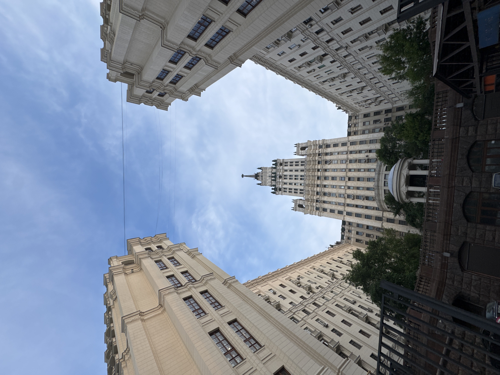 The photograph captures a low-angle view of the iconic Stalinist skyscraper, the Ministry of Foreign Affairs, located in Moscow, Russia. The grand building, with its distinctive tiered design and spire topped with a red star, dominates the center of the frame. Flanking the central tower are two identical, imposing residential buildings, also constructed in the Stalinist architectural style, their pale facades stretching upwards and inwards, creating a dramatic perspective. The buildings are adorned with numerous windows and decorative cornices. Between the flanking buildings and in front of the central tower, lush green trees are visible, adding a touch of nature to the urban landscape. A small, ornate rotunda with columns sits nestled in the foreground of the central structure. Below the buildings, a dark stone wall and a black metal fence are visible, suggesting a boundary or elevated promenade. The sky above is a soft blue, with scattered white clouds. The overall impression is one of monumental architecture against a serene sky.