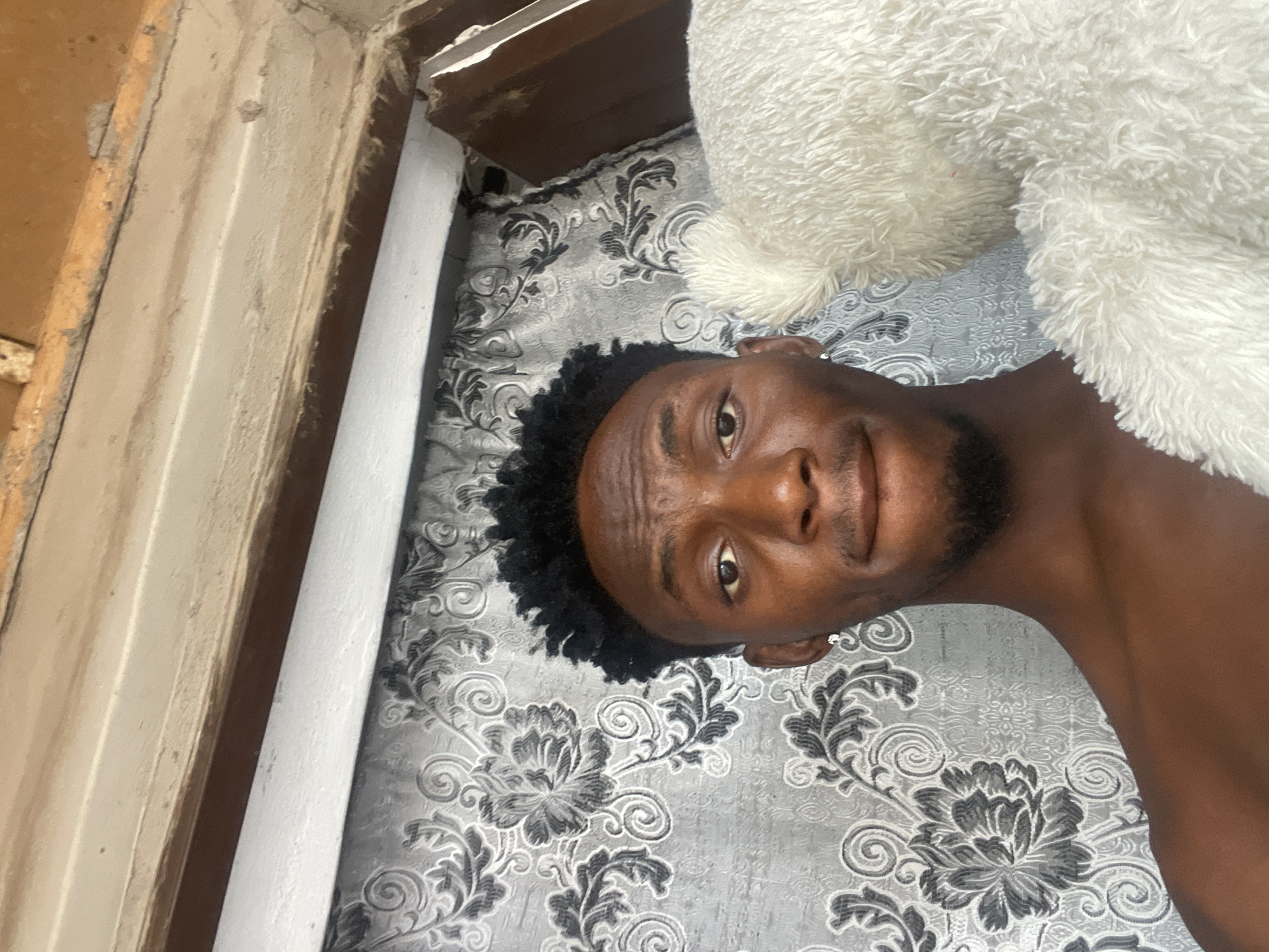 A medium close-up, portrait-style photograph depicts a young Black man lying on his back, looking directly at the camera with a neutral to slightly amused expression. The image is rotated 90 degrees clockwise, positioning his head on the left side of the frame and his body extending to the right.
The subject has dark skin, short, dark, curly hair, and a short beard or stubble. A small, subtle earring is visible in his left ear. He is shirtless. He appears to be relaxing indoors, possibly in a bedroom in Lokoja, Nigeria.
He is lying on a bed covered with a grey bedspread or sheet featuring a prominent white floral and scroll pattern. A large, fluffy white object, likely a pillow or blanket, partially covers the upper right portion of the frame, resting next to his head and upper body. To the far left of the image, parts of a light brown, possibly wooden, structure are visible, along with a white, pipe-like object running vertically. The lighting is soft and even, suggesting an indoor setting without direct sunlight. There is no visible text in the image.