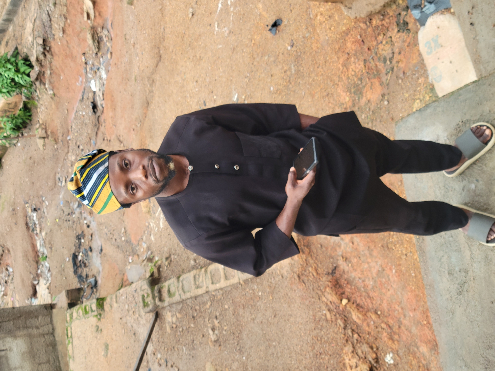 A man wearing a black traditional outfit and a multi-colored cap stands on a concrete surface outdoors. He is holding a smartphone in his right hand. The background shows a dirt path with some vegetation and scattered debris. The overall setting appears to be an urban or semi-urban area.