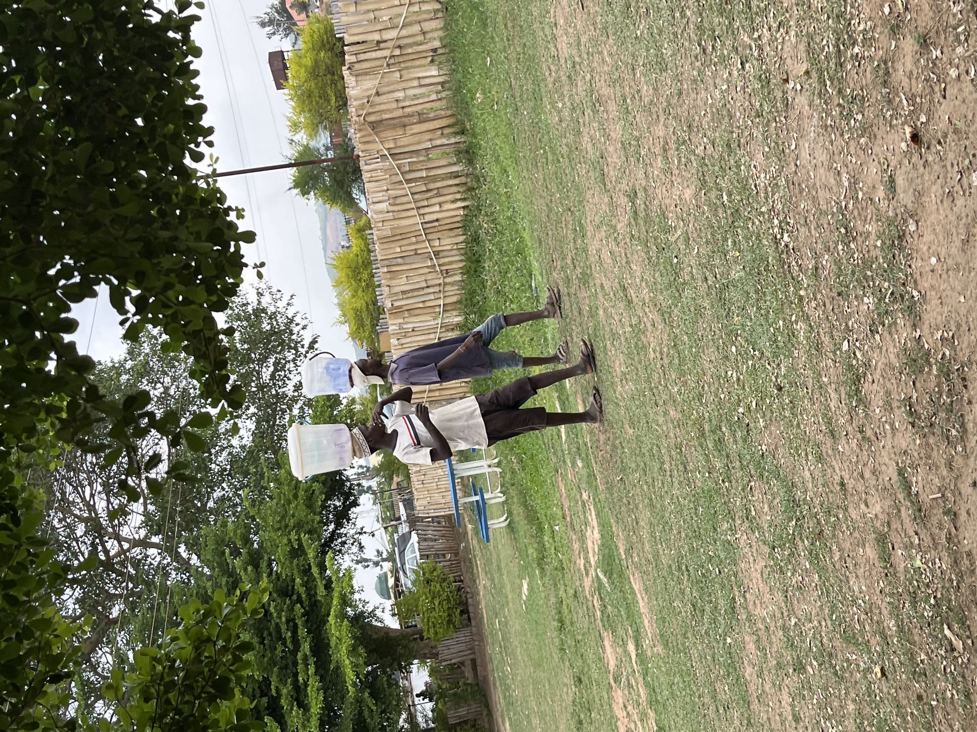 Two young men are walking across a grassy field in Galadimawa, Nigeria. Each is carrying a rectangular plastic container on their head. The man in the foreground is wearing a white t-shirt, dark shorts, and sandals. The man behind him is wearing a dark shirt, shorts, and sandals. A bamboo fence borders the field. Trees surround the location.