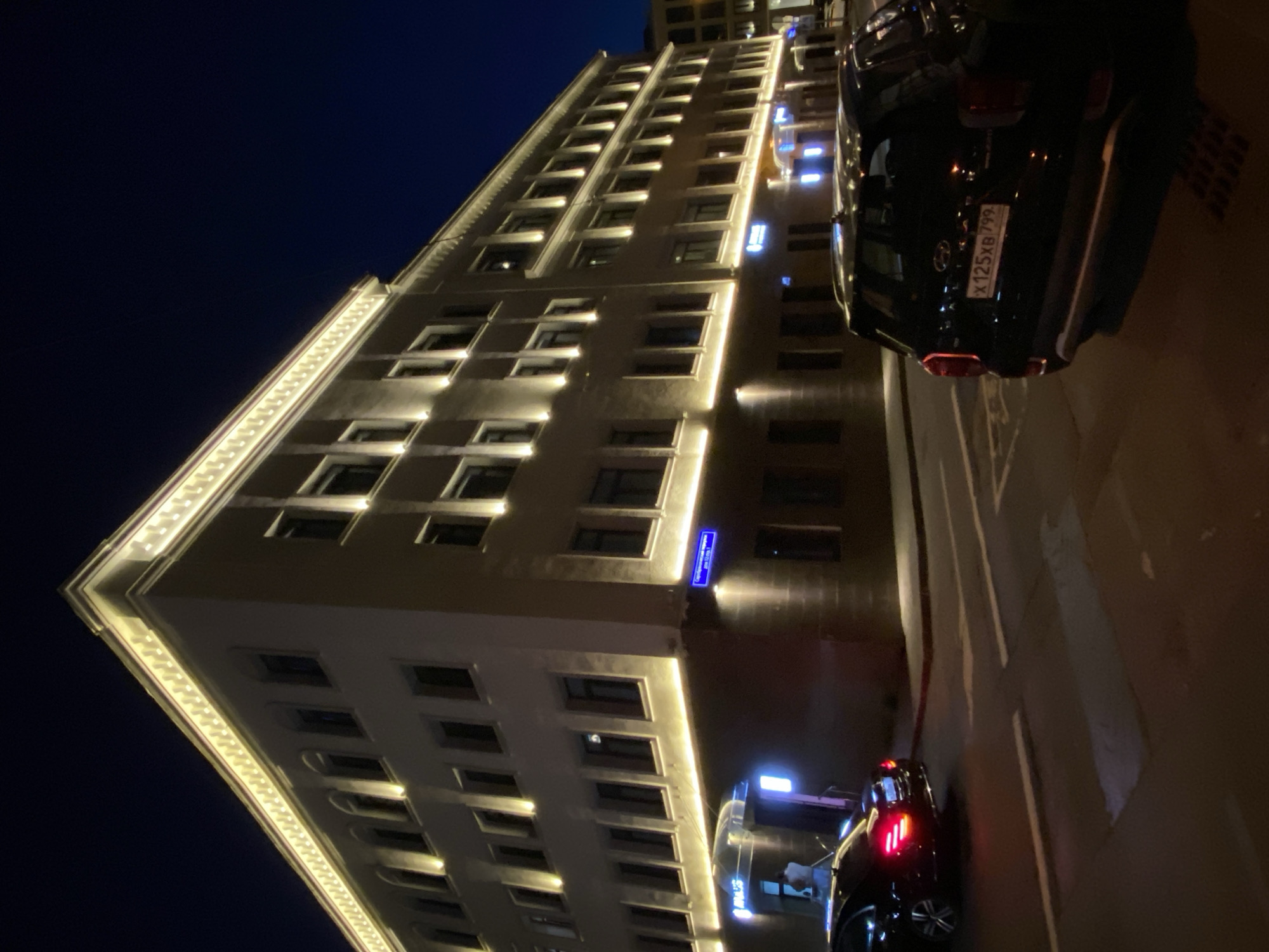 Captured at night, the image presents a view of a multi-story building with illuminated edges, situated in Moscow, Russia. The building has numerous windows and architectural details, including a lit cornice. In the foreground, two parked cars are visible. The registration plate of the car closest to the viewer displays the Cyrillic characters "X 125 XB 799." A street sign can also be seen on the building's facade. The overall scene portrays an urban setting at night.