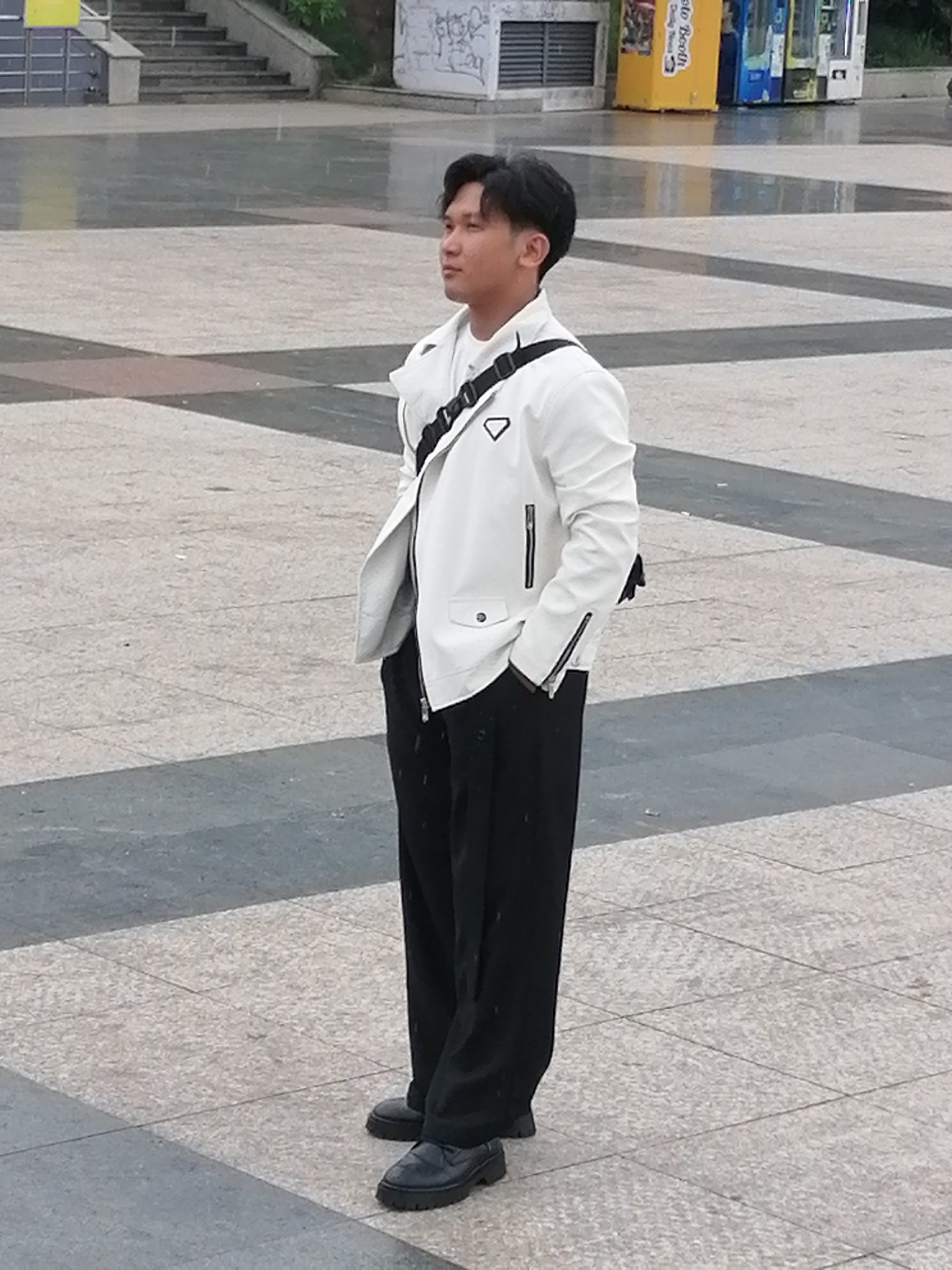 A young Asian man stands in a wide, paved plaza, looking to his right. He is wearing a white biker jacket with a black strap across his chest, black wide-leg pants, and black loafers. The plaza has a geometric pattern of light and dark tiles. In the background, there are concrete steps leading up to a building with graffiti, and some brightly colored vending machines. The ground appears wet, suggesting it may have rained recently. The overall setting is an urban public space.