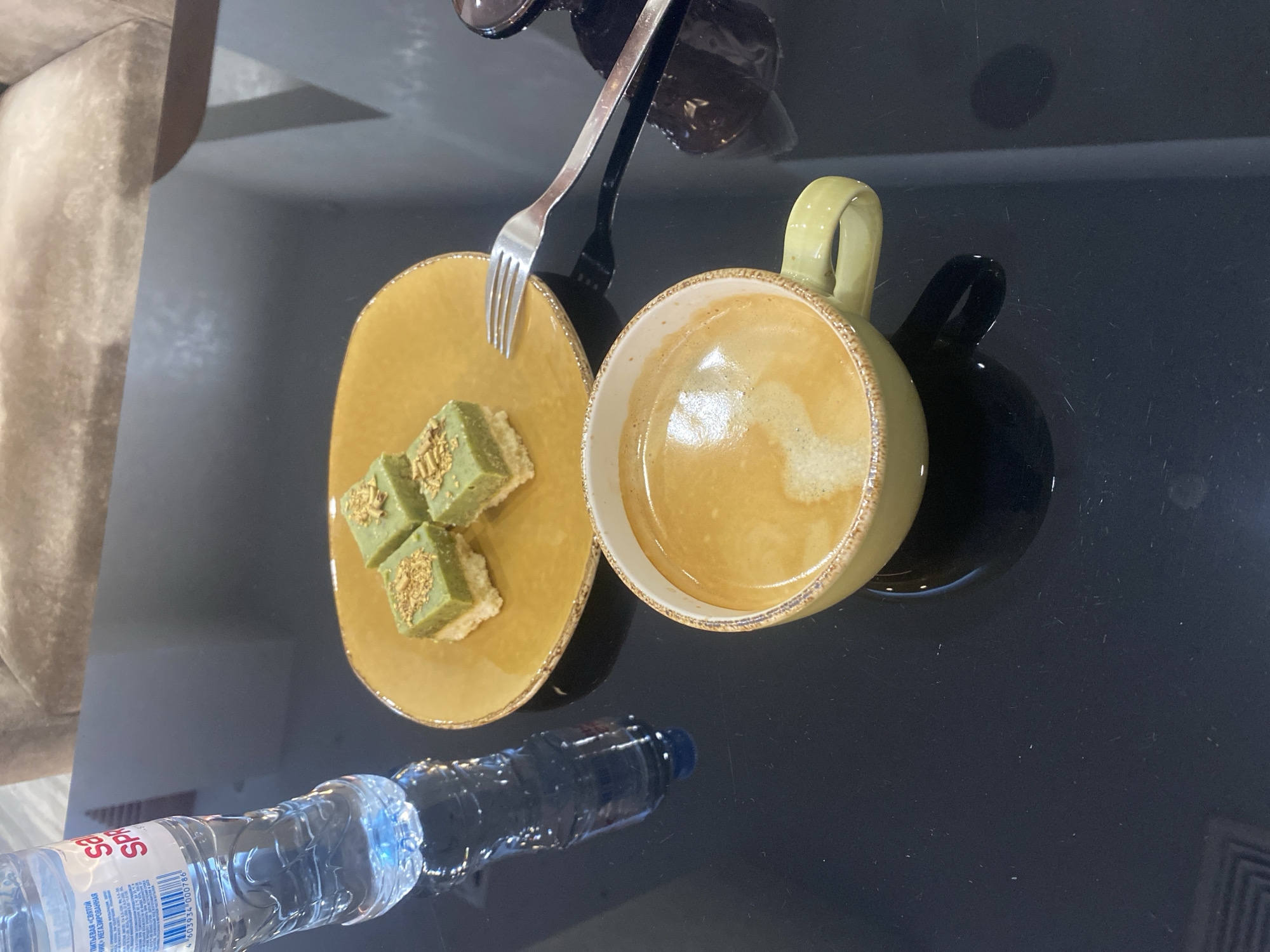 The image shows a table with a bottle of water, a plate of pastries, and a cup of coffee. The pastries are small and square, topped with green frosting and crushed nuts. The coffee is in a light green mug. A fork is placed on the table next to the plate. The table is black and reflective, suggesting it might be made of glass or a similar polished material. In the background, a beige upholstered sofa is visible, indicating an indoor setting, possibly a cafe or lounge area. The lighting is soft, casting subtle shadows. The overall scene suggests a moment of relaxation or a break. The presence of a specific bottle of water, "S.Pellegrino," could be a clue to the location. S.Pellegrino is a popular brand of sparkling water often found in cafes and restaurants internationally. Without further visual cues specific to Minsk, Belarus, a precise location within the city cannot be determined from the image alone. However, the setting is consistent with an urban cafe or lounge environment that might be found in Minsk.