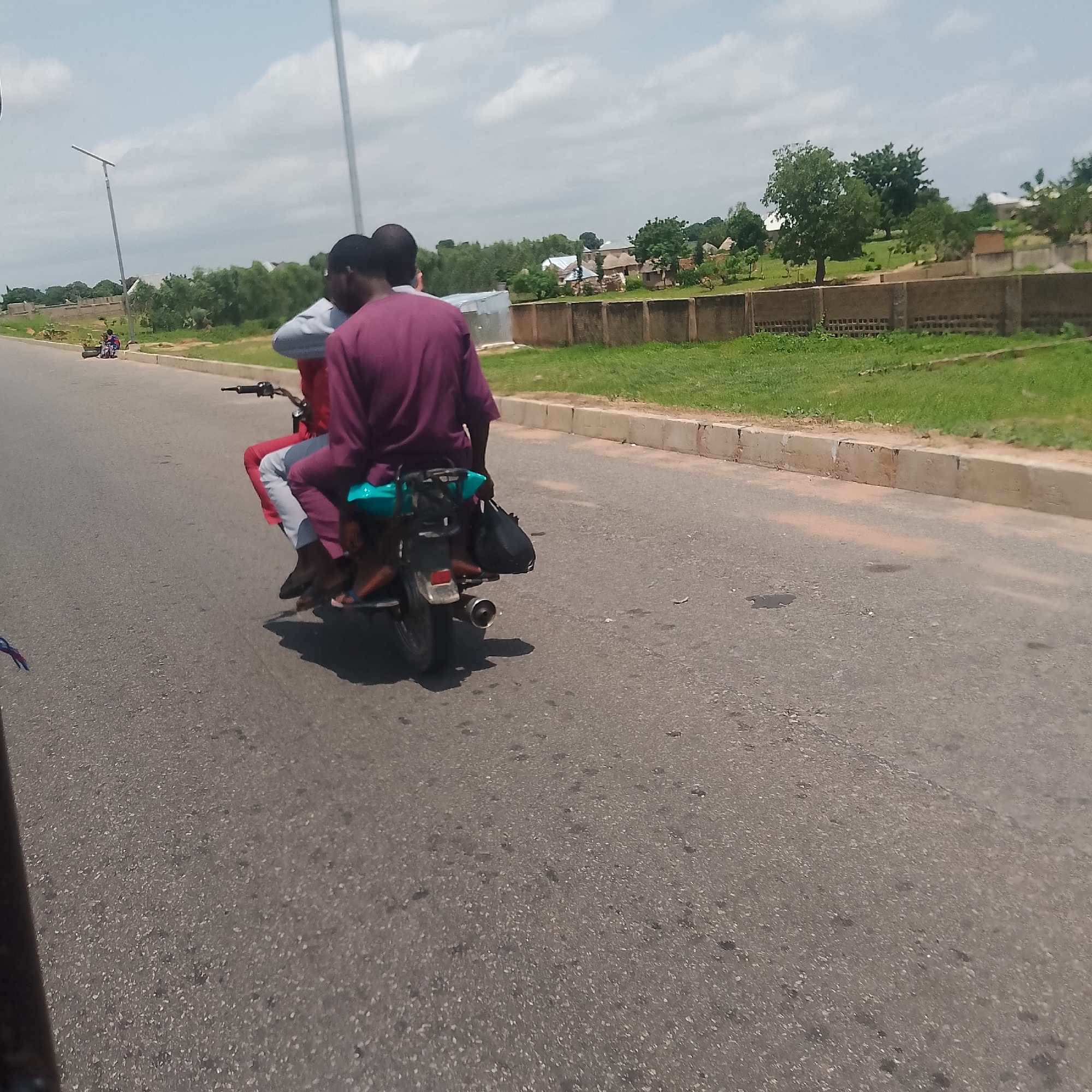 In Jalingo, Nigeria, on a paved road with a concrete curb and a grassy shoulder, two individuals are riding on a motorcycle. The person in the rear, wearing a maroon robe, is operating the motorcycle. The person in front is wearing a light blue garment and maroon pants. The motorcycle has a turquoise seat and a black bag attached to the rear. In the background, there are buildings with thatched roofs and trees. Further in the distance, a street light is visible. The sky is cloudy.