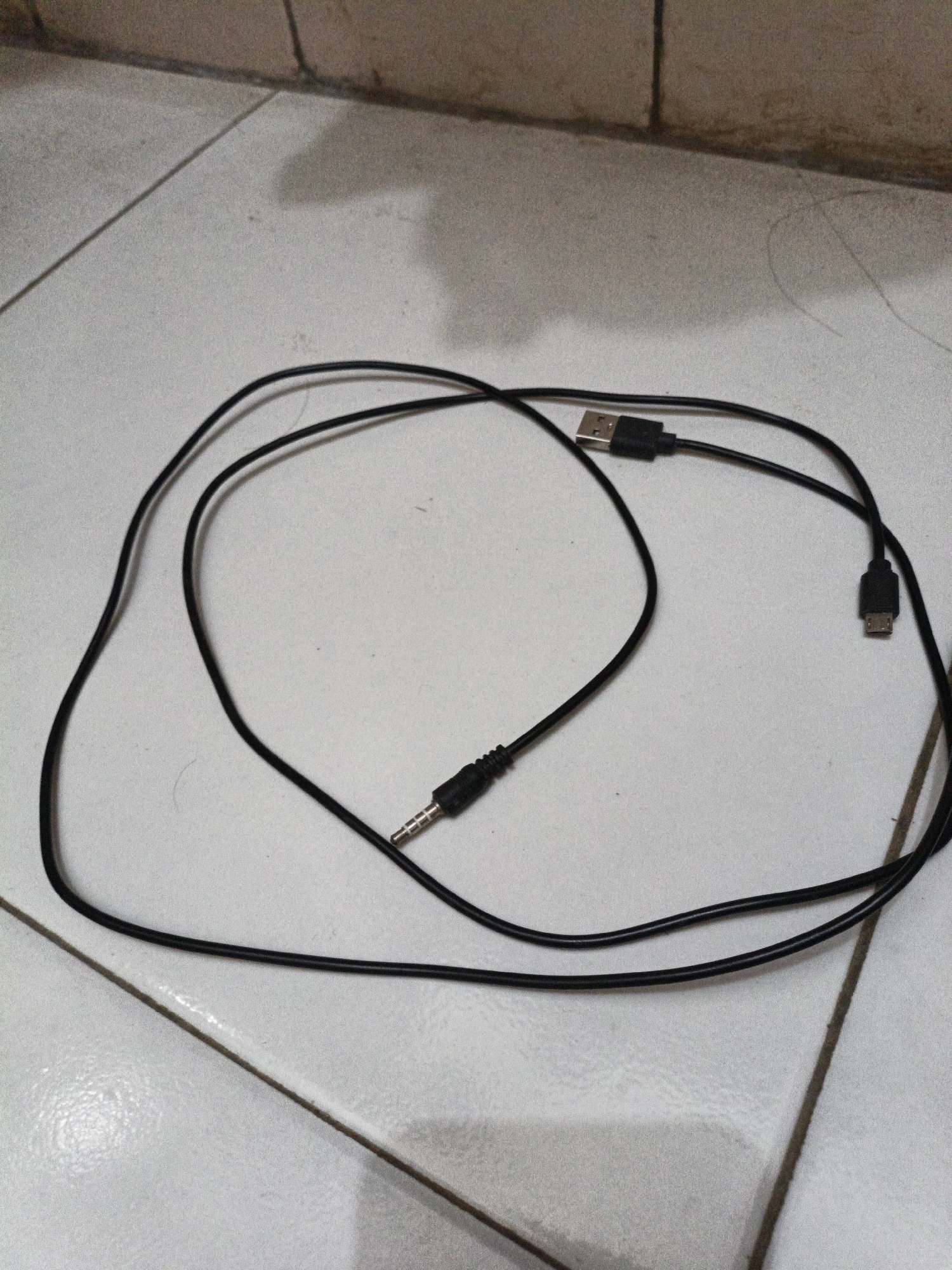 A black charging cable with a USB connector and a micro USB connector is displayed on a tiled floor. The cable is partially coiled and appears to be in use or recently used. The background consists of a tiled wall and floor. The scene is indoors, in an unspecified location, and no specific details about Cilegon, Indonesia are visible. There are no people or animals present.