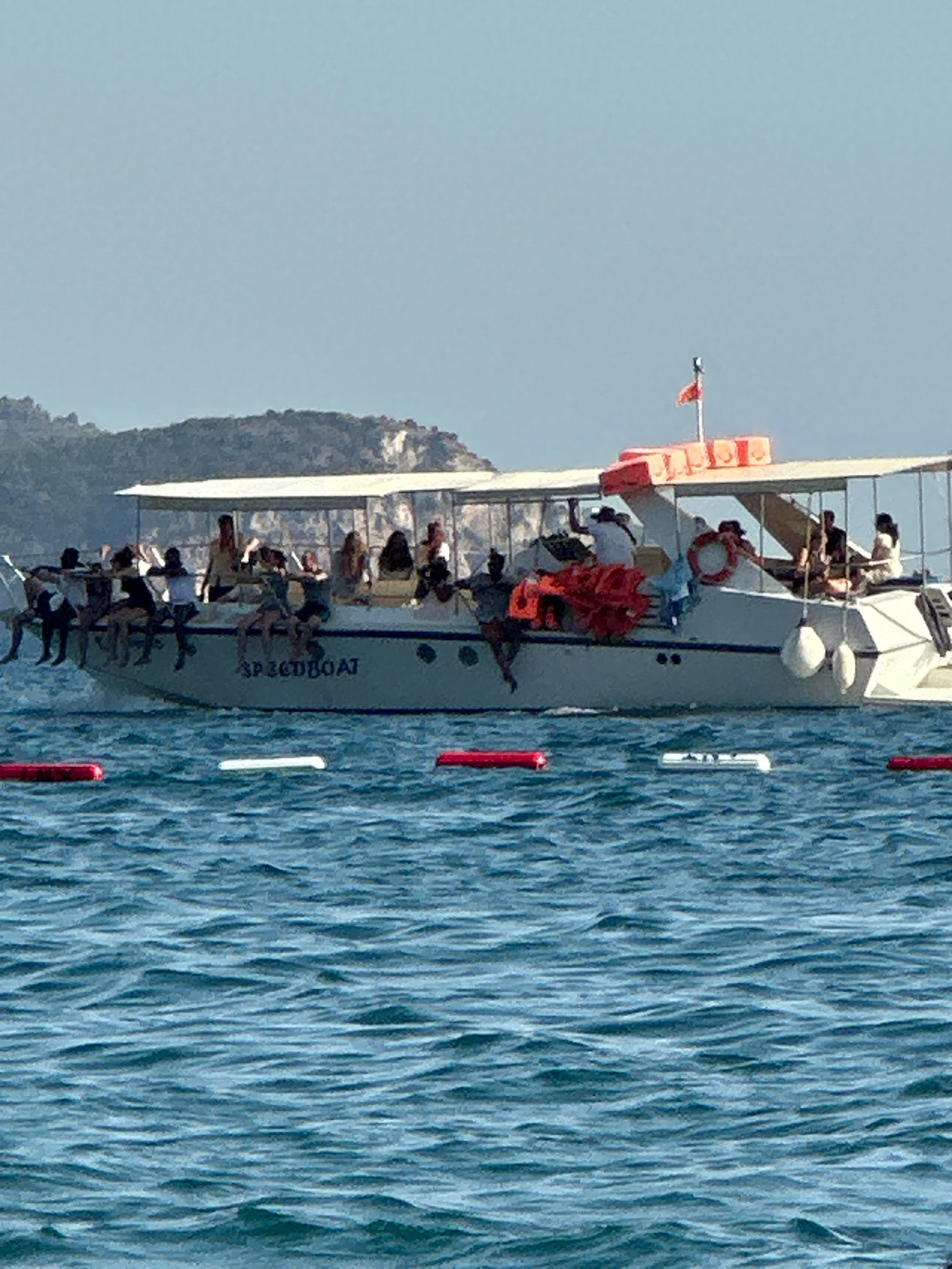 A speedboat named "SPEEDBOAT" is traveling on blue, rippling water under a clear, pale blue sky. The boat is carrying approximately 20 people, some of whom are sitting on the edge of the boat, with their legs dangling in the water. The boat has a white hull with a dark blue stripe, and a white canopy covering the upper deck. There are several orange life jackets on the deck of the boat, and a life ring is visible on the side. In the background, a tree-covered island with rocky outcrops rises from the water. The scene appears to be in a coastal area, possibly a tourist destination.