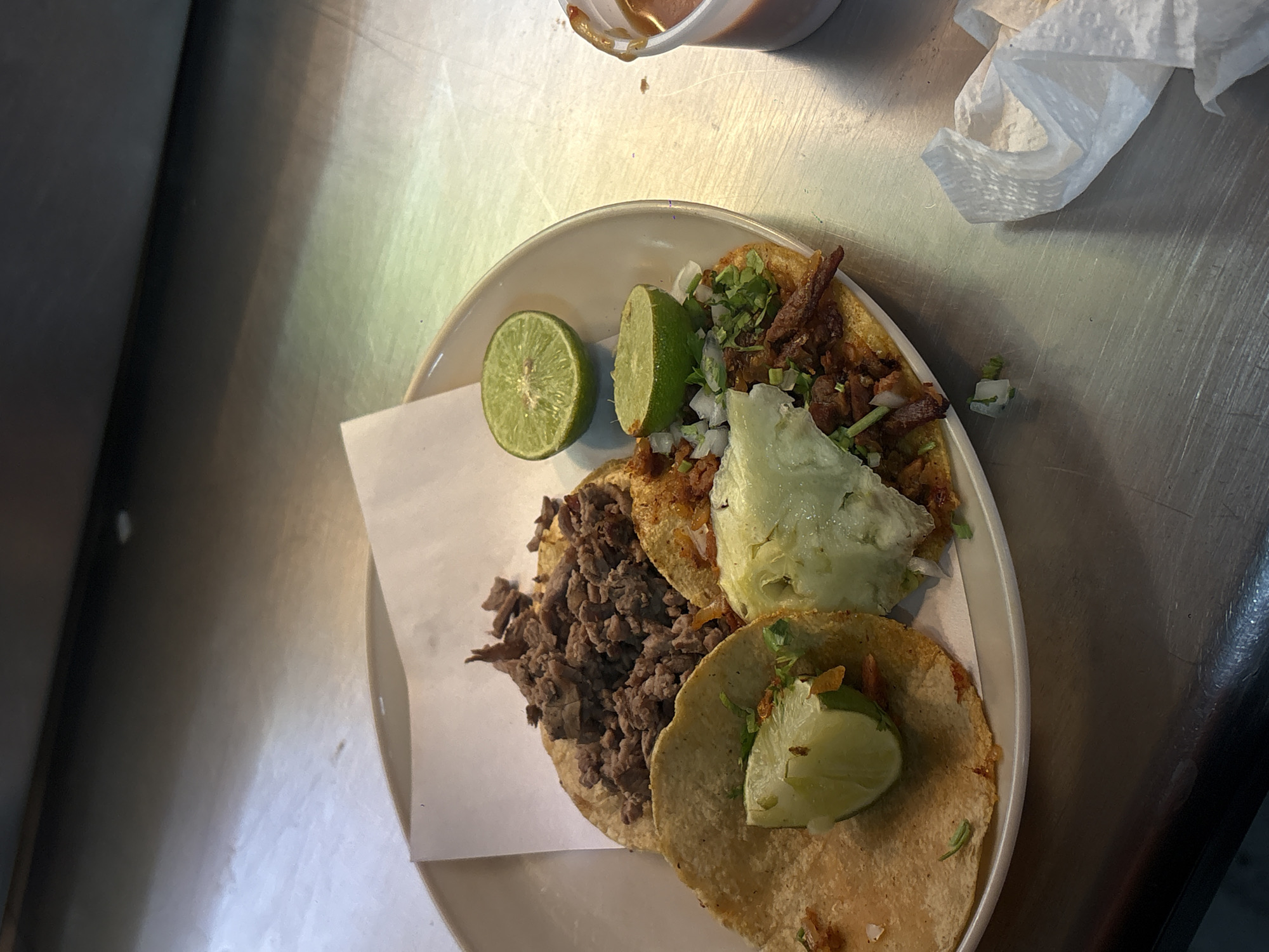 A plate of three tacos is shown on a metal surface. The tacos appear to be made with corn tortillas and are filled with different meats and toppings. One taco has chopped meat with onions, and it is garnished with a wedge of lime. Another taco is filled with a darker, shredded meat, also topped with a lime wedge. The third taco contains meat with cilantro and what appears to be lettuce or cabbage. A half of a lime is placed between two of the tacos. At the top of the frame, there is a small container of sauce and a crumpled white napkin. The setting appears to be casual, possibly a taqueria in Mexico City, Mexico.
