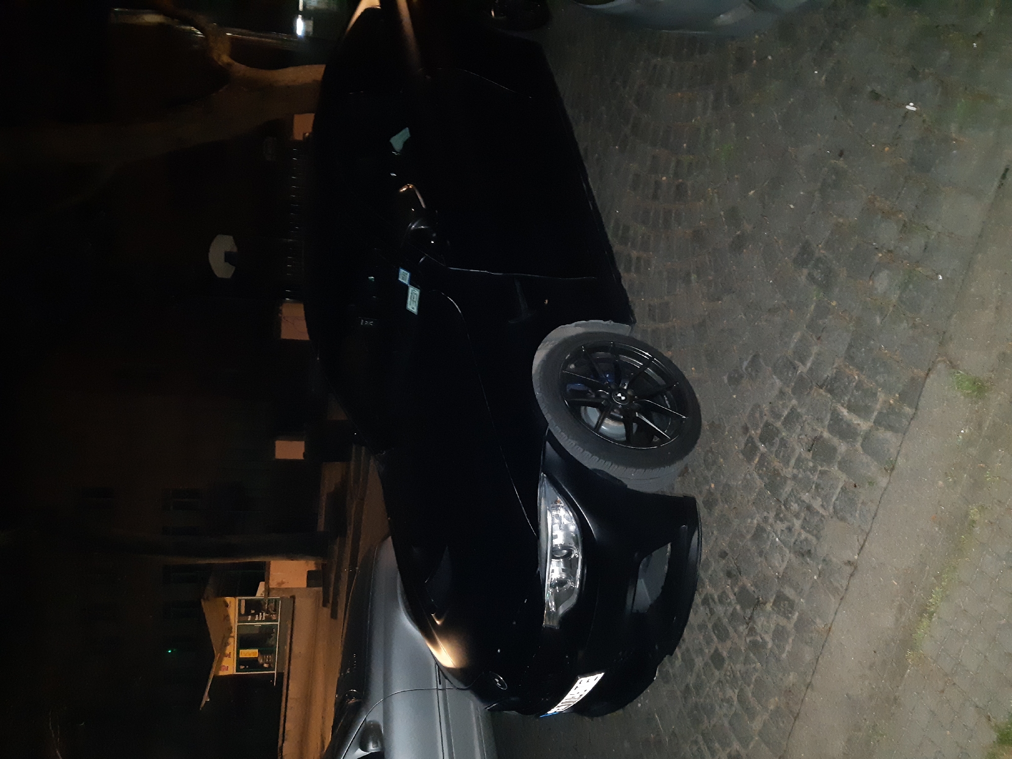 A nighttime view depicts a black car parked on a cobblestone street in an urban area. The lighting is dim. The car's front right side is visible, including a wheel with black rims. The vehicle is positioned alongside a building or structure in the background. No people are visible. The location is presumed to be Plovdiv, Bulgaria.