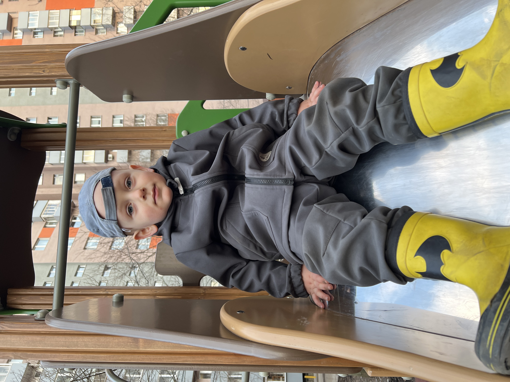 A child, dressed in a gray tracksuit and yellow Batman-themed rain boots, is lying on a metal slide in a playground. He wears a gray cap. The playground is located near a multi-story building in Mytishchi, Russia. Bare tree branches are visible in the background, suggesting it is late autumn or early spring. The playground structure features brown and green elements.